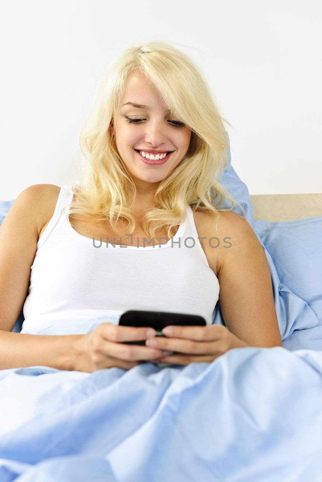 Smiling blonde woman texting on phone in bed at home