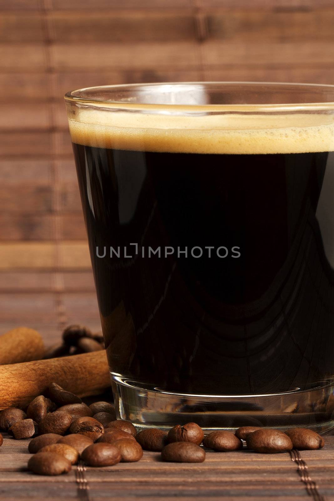 espresso in a short glass with coffee beans and cinnamon sticks from side on wooden background
