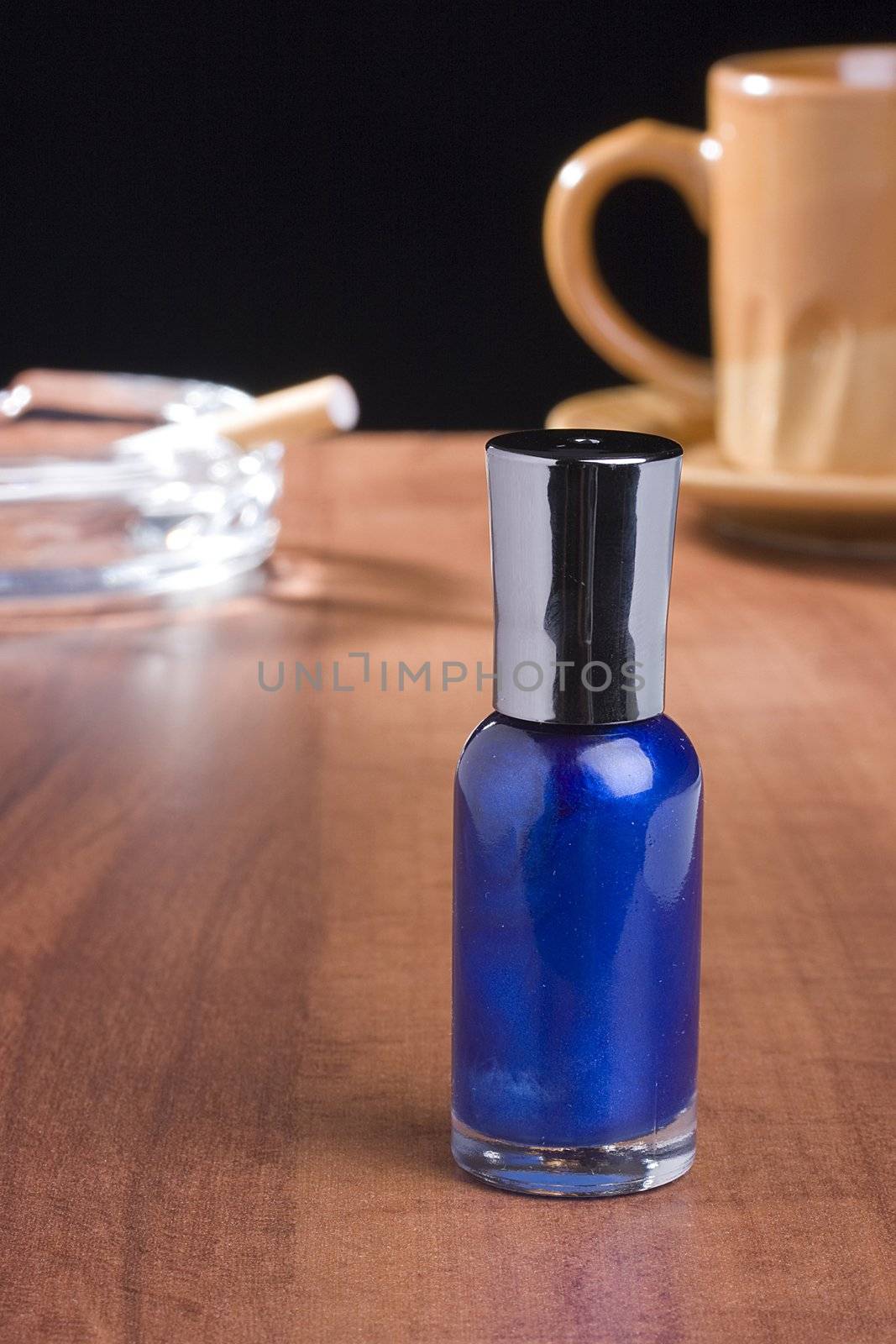 Blue nail polish standing on a coffee table in front of a cup of coffee and an ashtray.