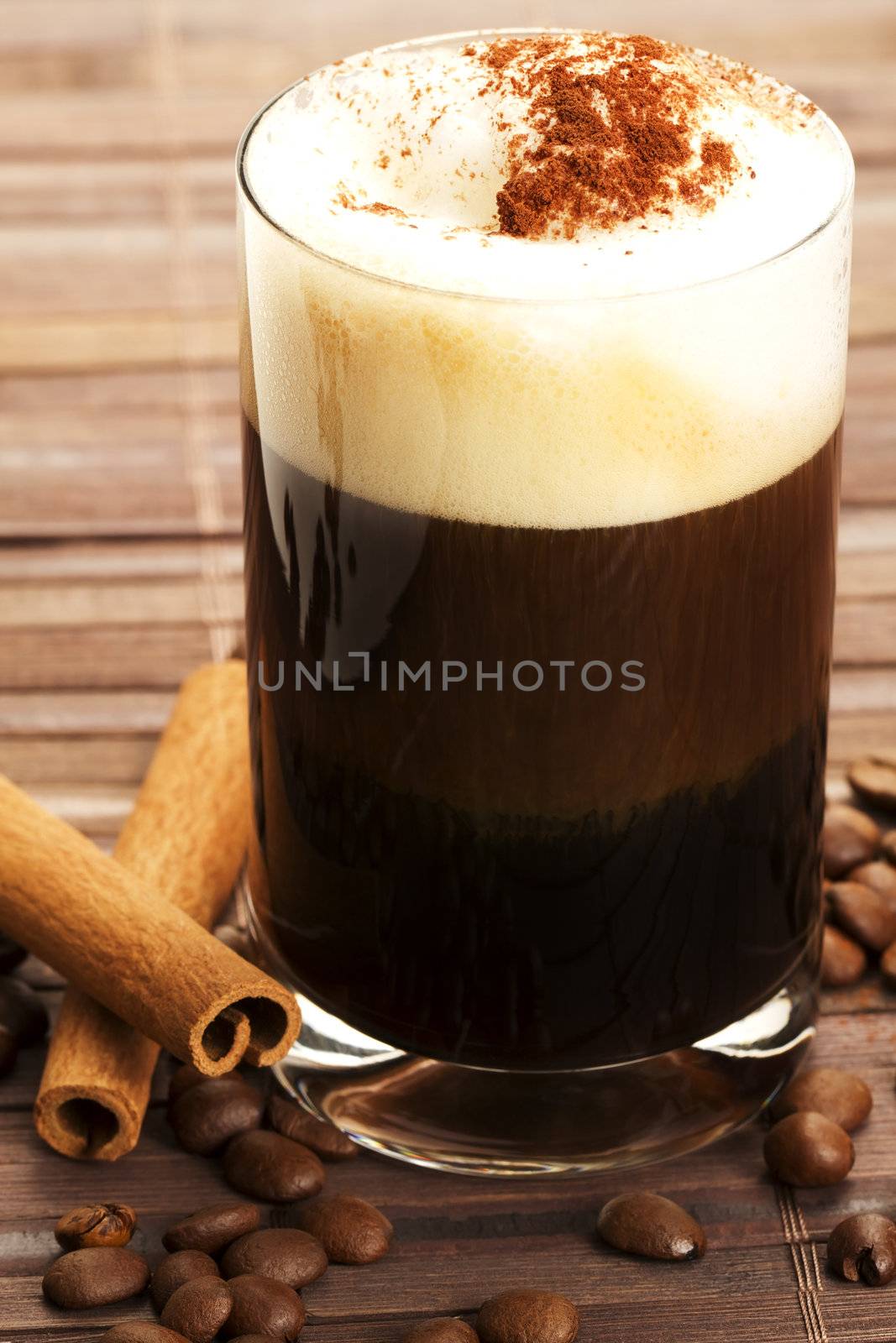 espresso in a straigt glass with milk froth cocoa powder, cinnamon sticks and coffee beans aside on wooden background