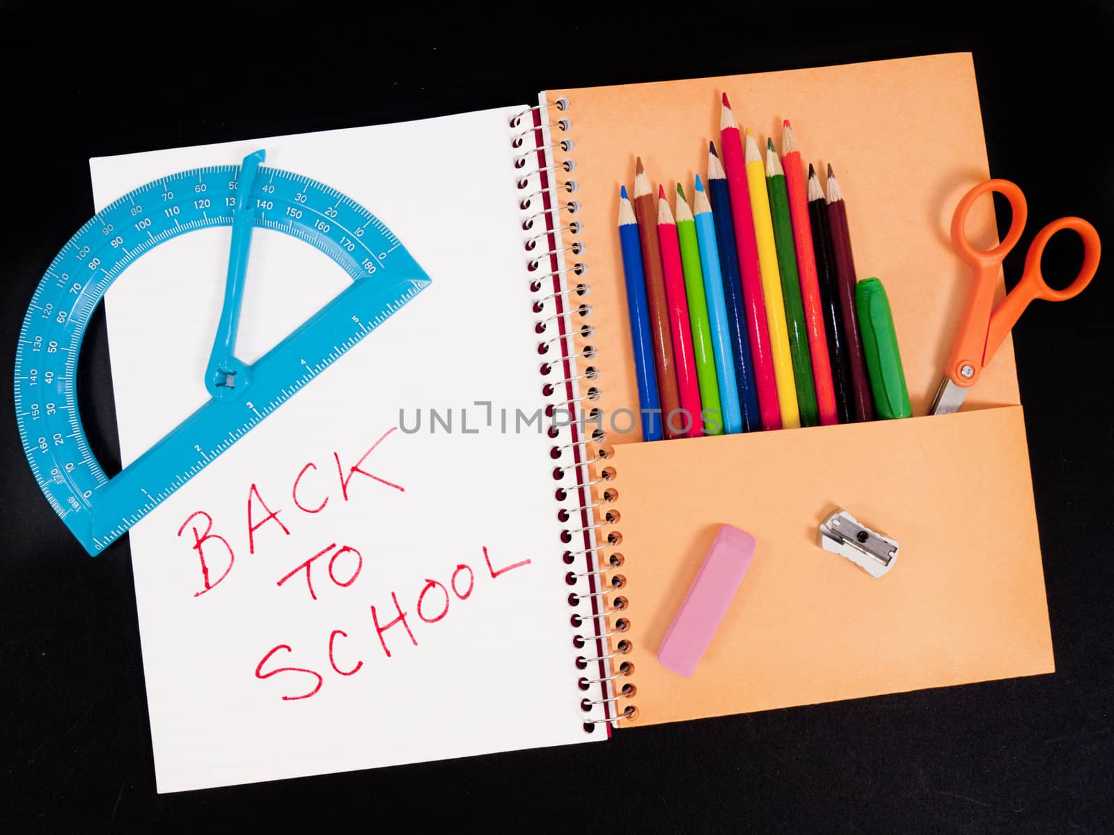 Back-to-school theme of student notebook with pocket full of colored pencils. Other items are scissors, protractor, sharpener, and eraser. Background is black.