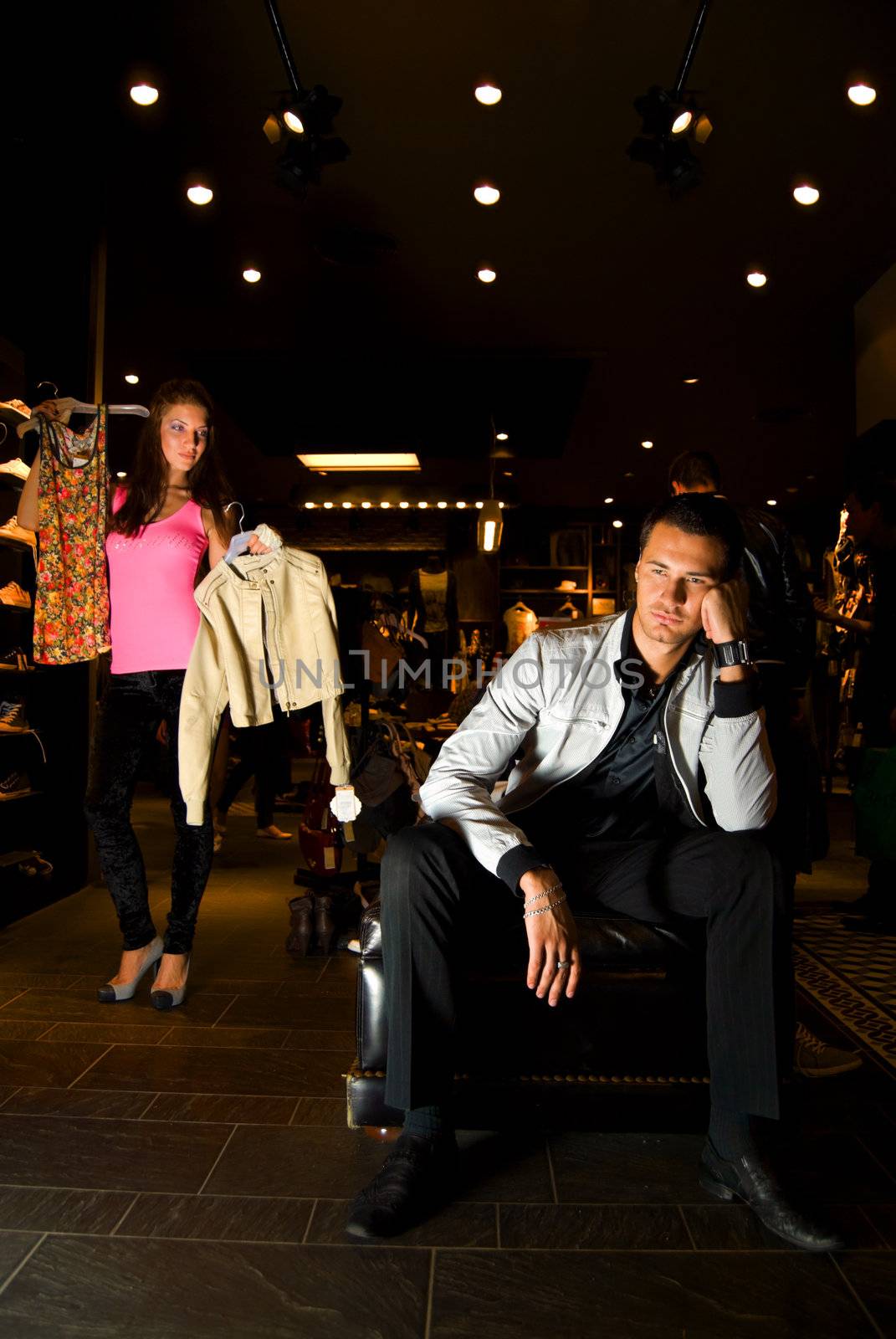 Pair of young adults trying on new clothes in dress storee. The man in fashion suit tired of infinite woman's clothes. His girl friend shows him different variants of wearings.