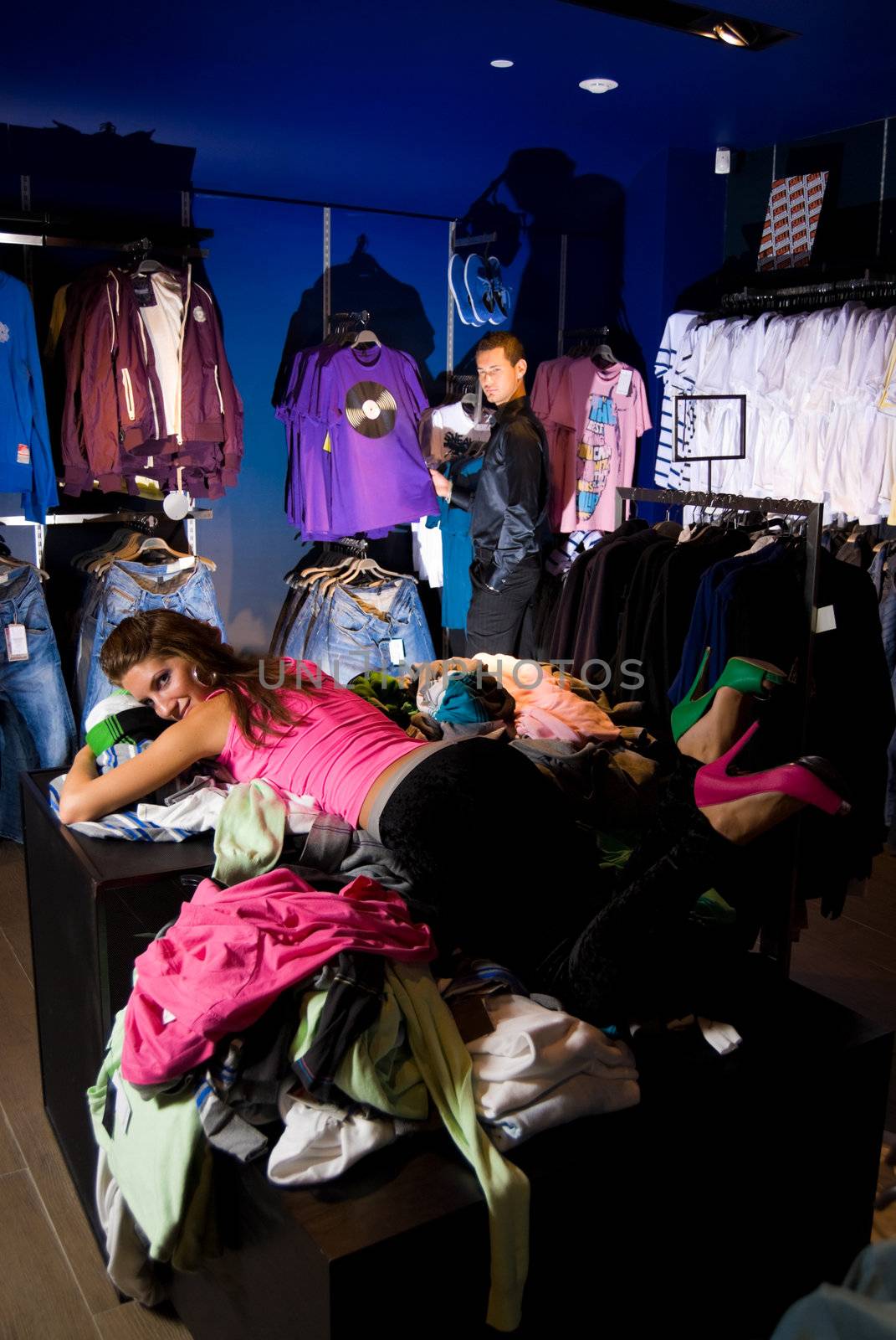 Pair of young adults in dress store. Girl lays on pile of shirts
