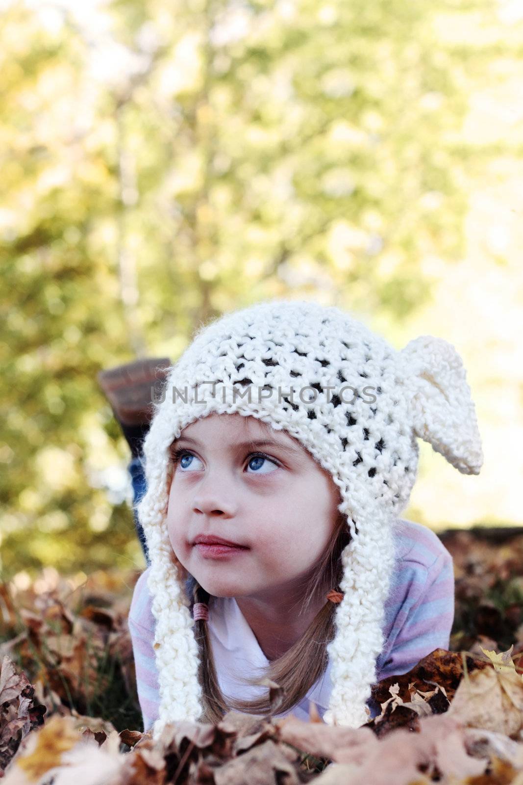 Beautiful little girl in a white bobble hat lying in the leaves and looking away from the camera. Selective focus on little girls face with shallow DOF. 
