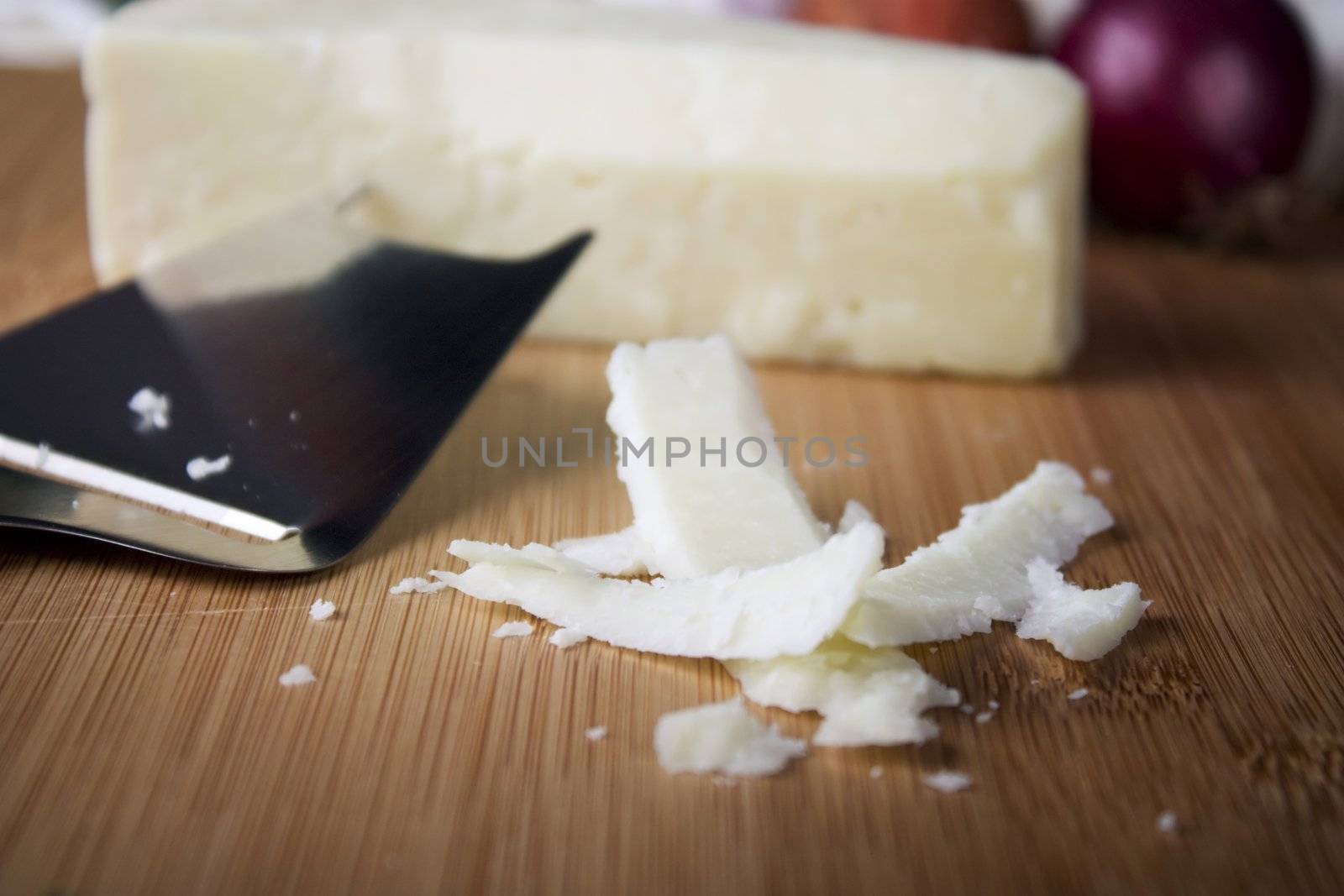 Flakes of italian cheese on cutting board with cheese slicer and block of cheese in background.  Shallow depth of field.