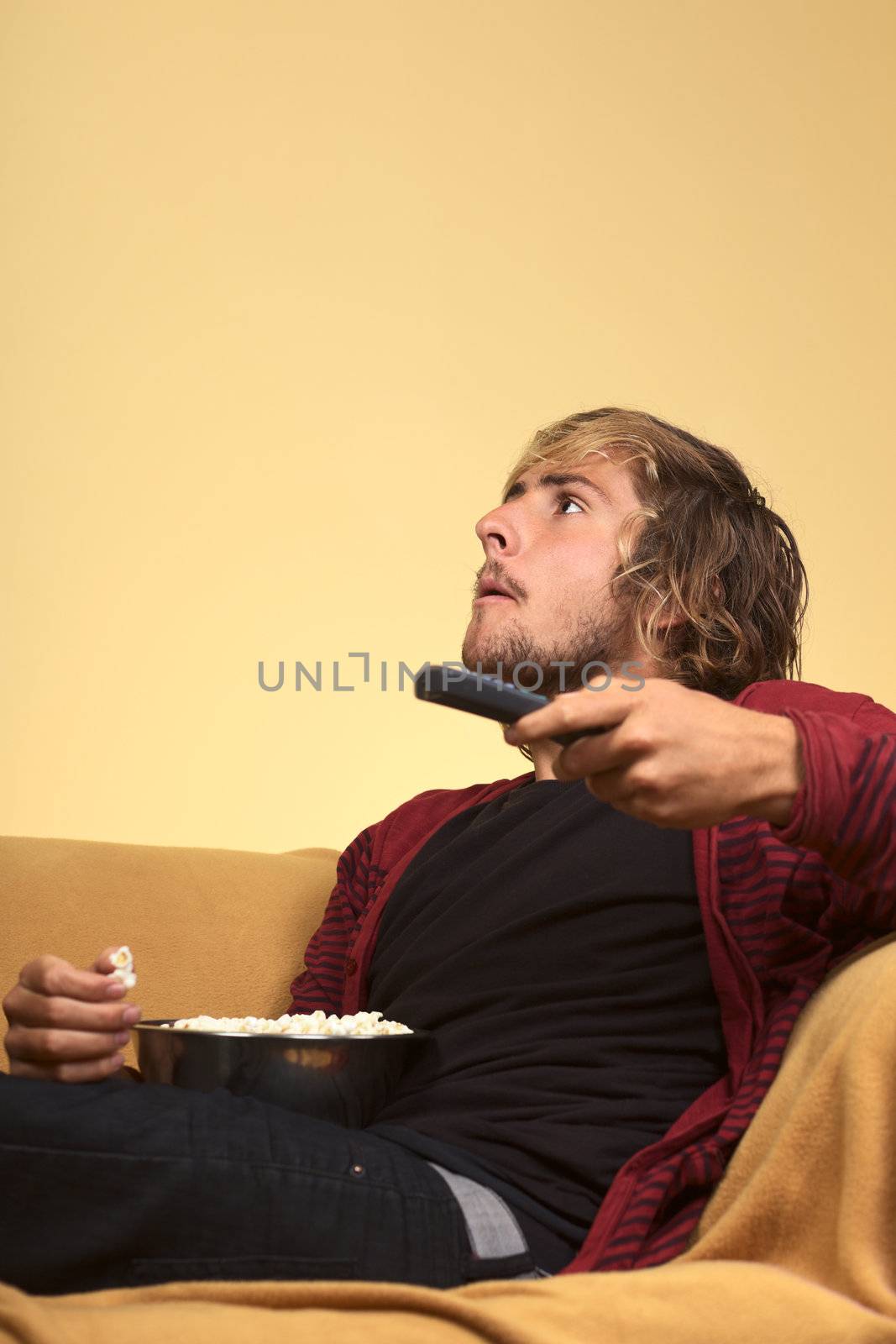 Young man watching TV holding a remote control in one hand and eating popcorn (Selective Focus, Focus on the left eye)