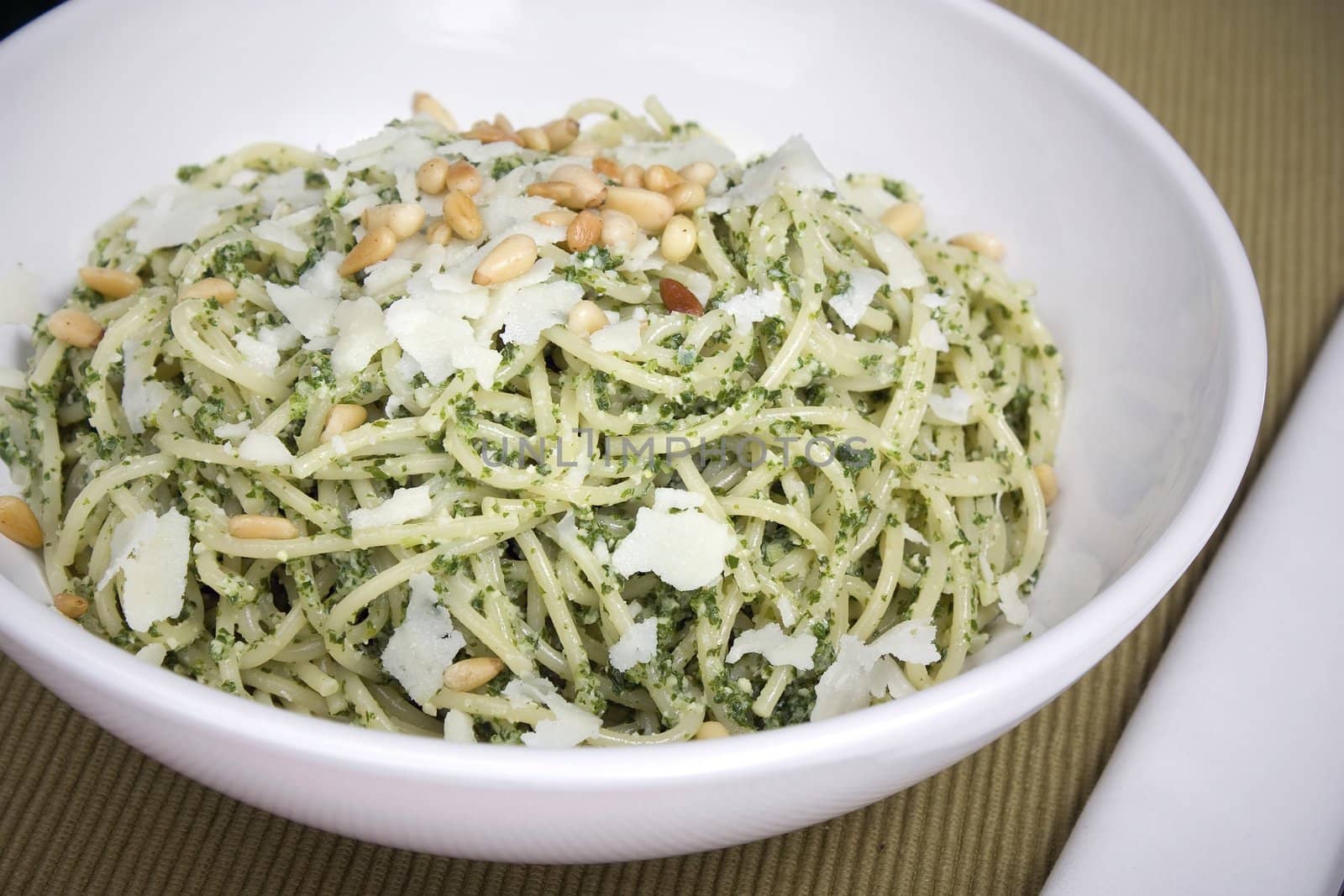 Pesto pasta in white bowl topped with Parmesan flakes and roasted pine nuts.