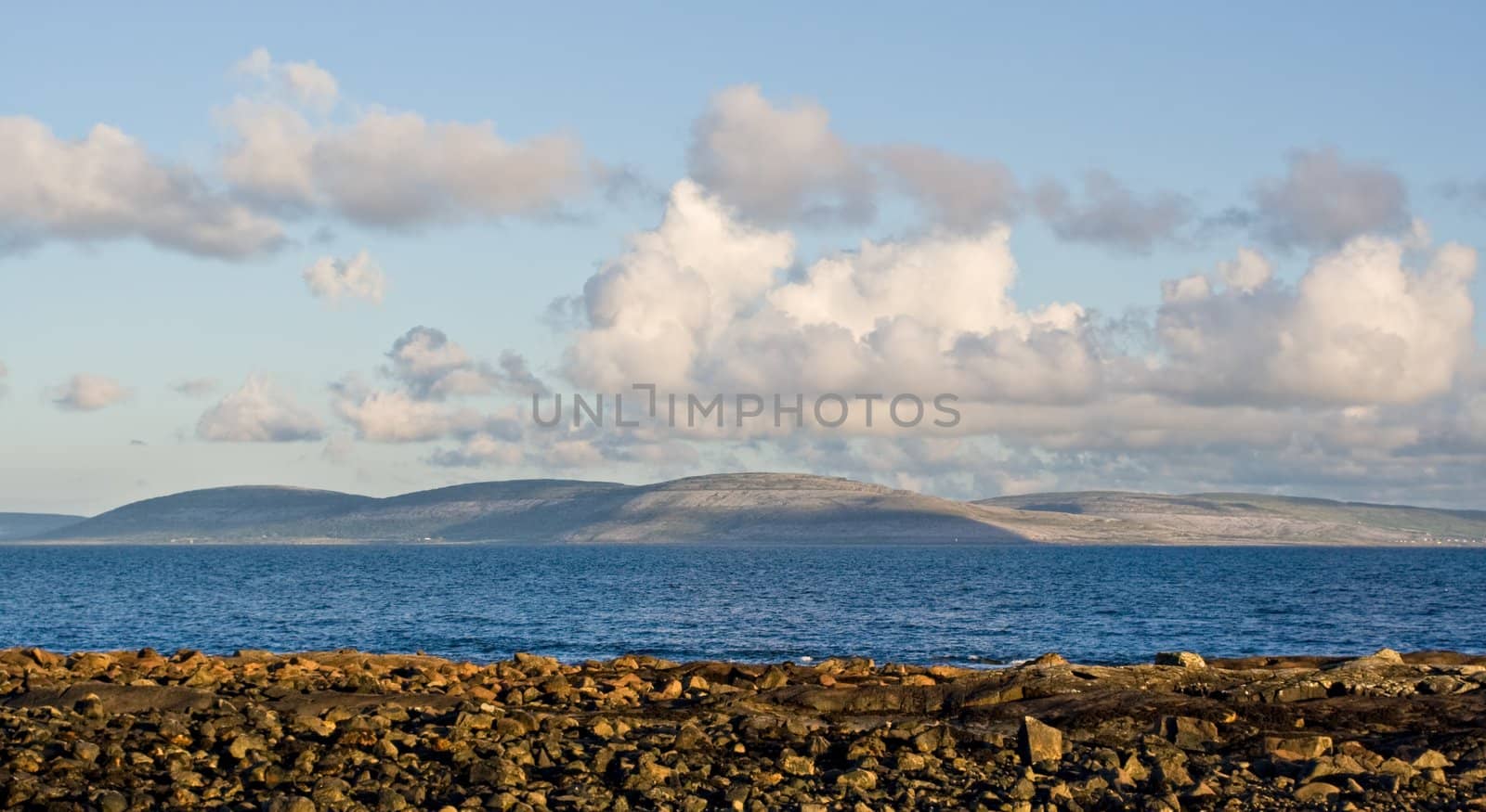 Galway Bay in Ireland from the town of Spiddal with The Burren across the bay. Photo is layered from front to back with Rocks, Galway Bay, The Burren, and sky.