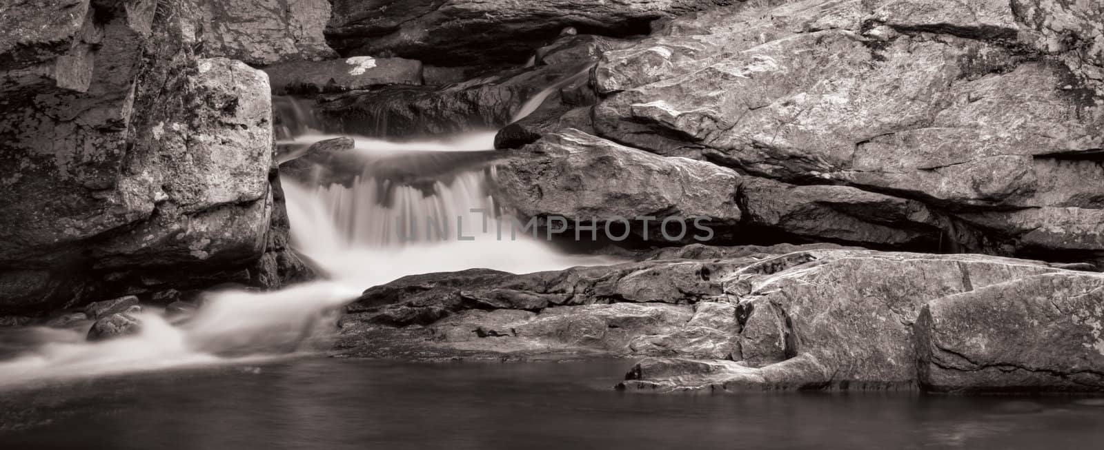 A small waterfall over rock in panorama format. Photo is in black and white.