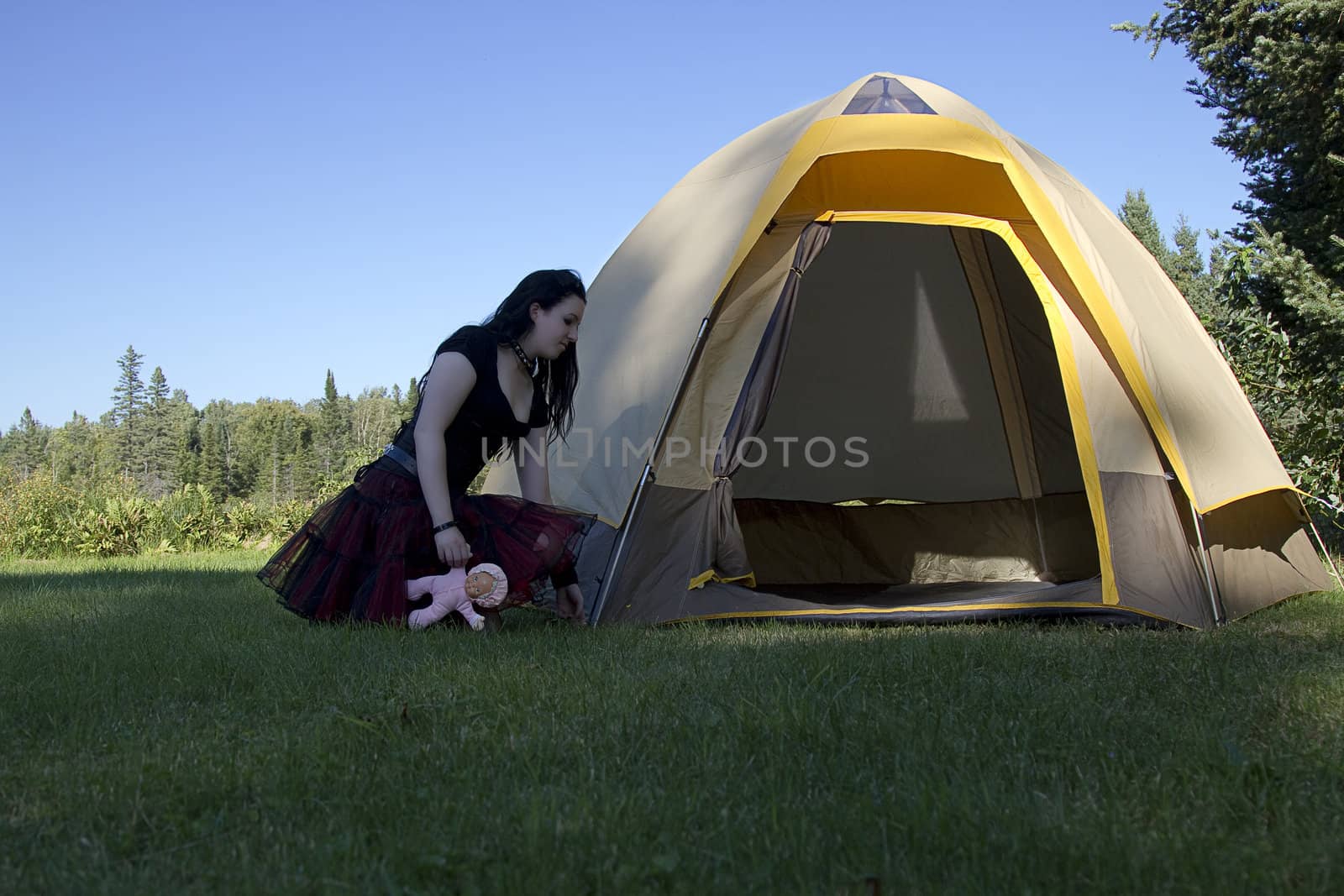 twenty something women wearing goth style clothes and holding a small baby doll adjusting a camping tent