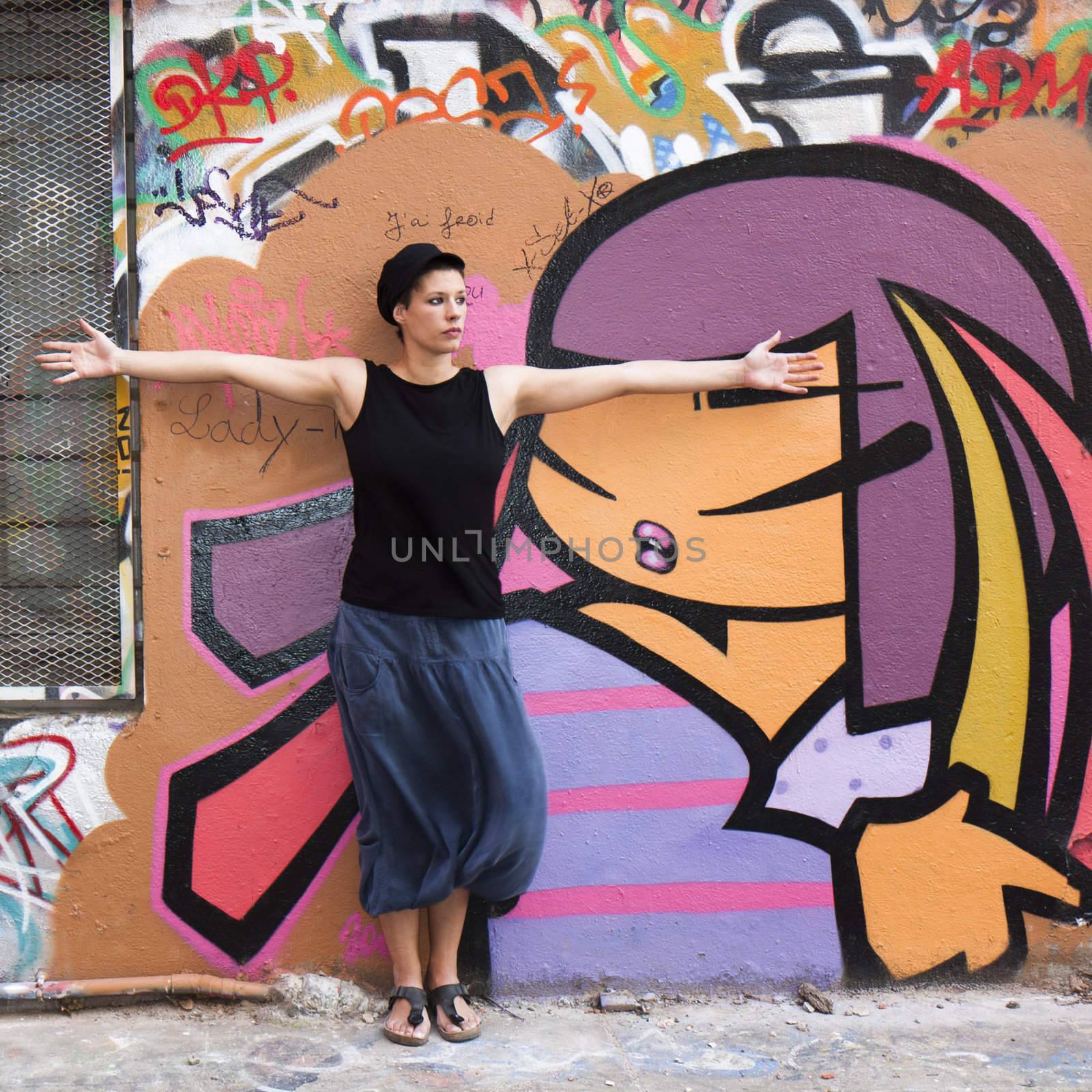 A young girl stands before a wall covered with graffiti, arms outstretched