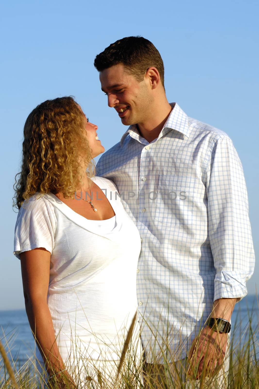A young happy couple is standing together at the beach with the sea in the background.