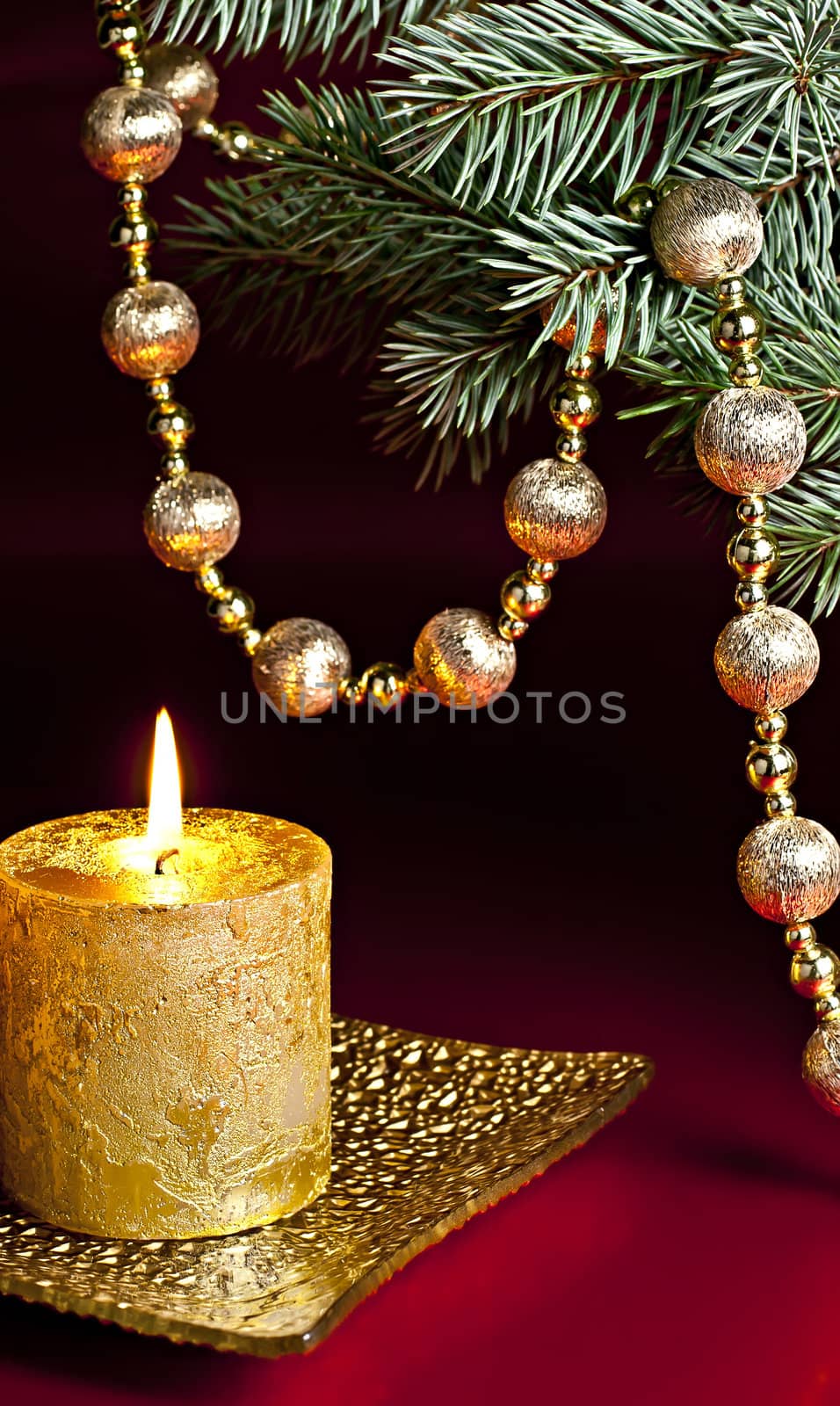 On a red background, a burning candle, fir branch and a ball chain.
