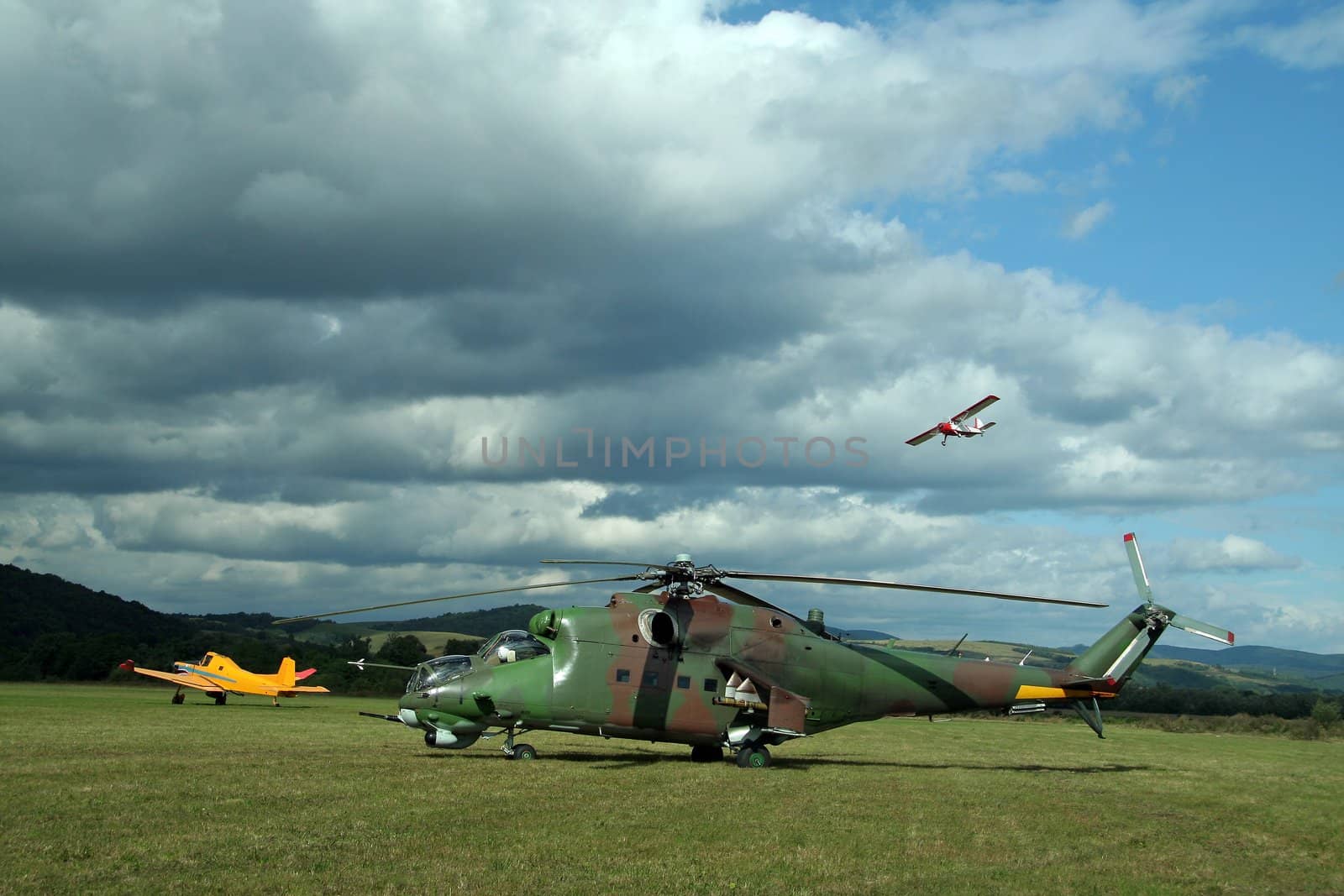 army helicopter and yellow plane on green airfield, white airplane in air, heavy clouds 