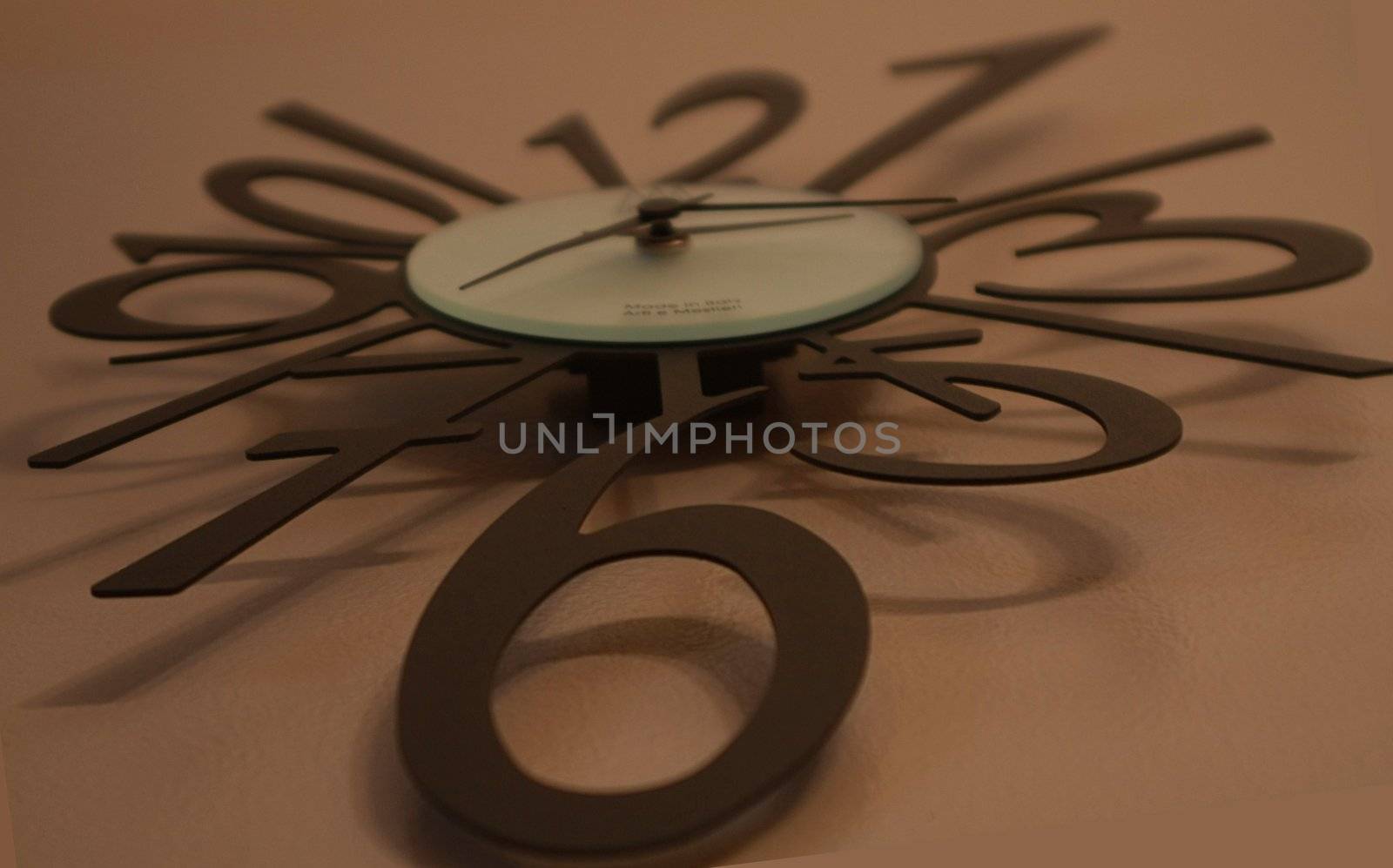 Large abstract wall clock with modern black numbers and hands. Clock is resting against a tan background.