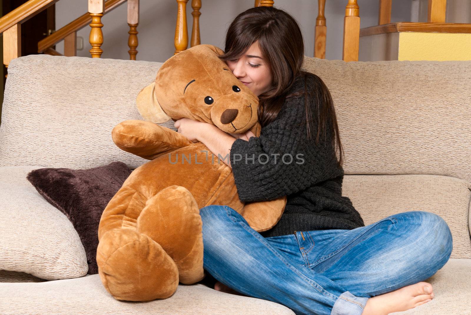 Young woman embracing teddy bear sitting on sofa by dgmata