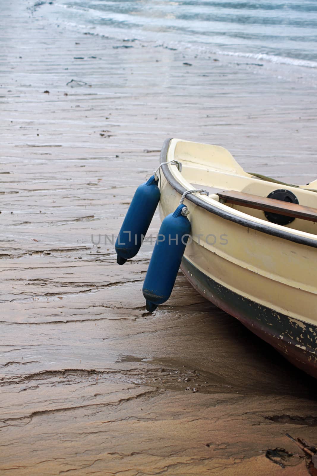 A cream coloured row boat with two blue fenders hanging from its side. Set on a sandy beech with the sea visible to top  of image.