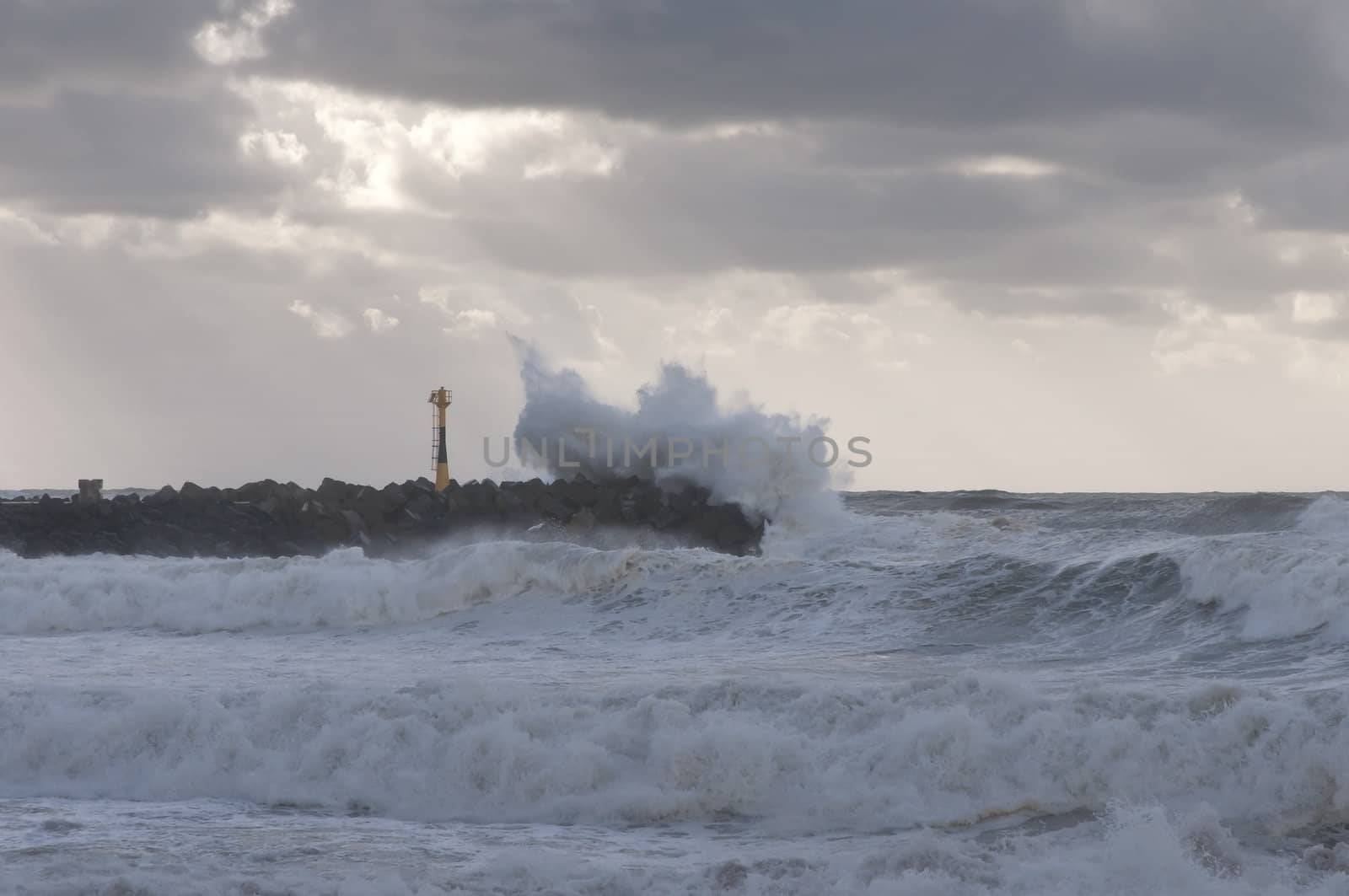 Big wave against a jetty with a little lighthouse during a stormy day
