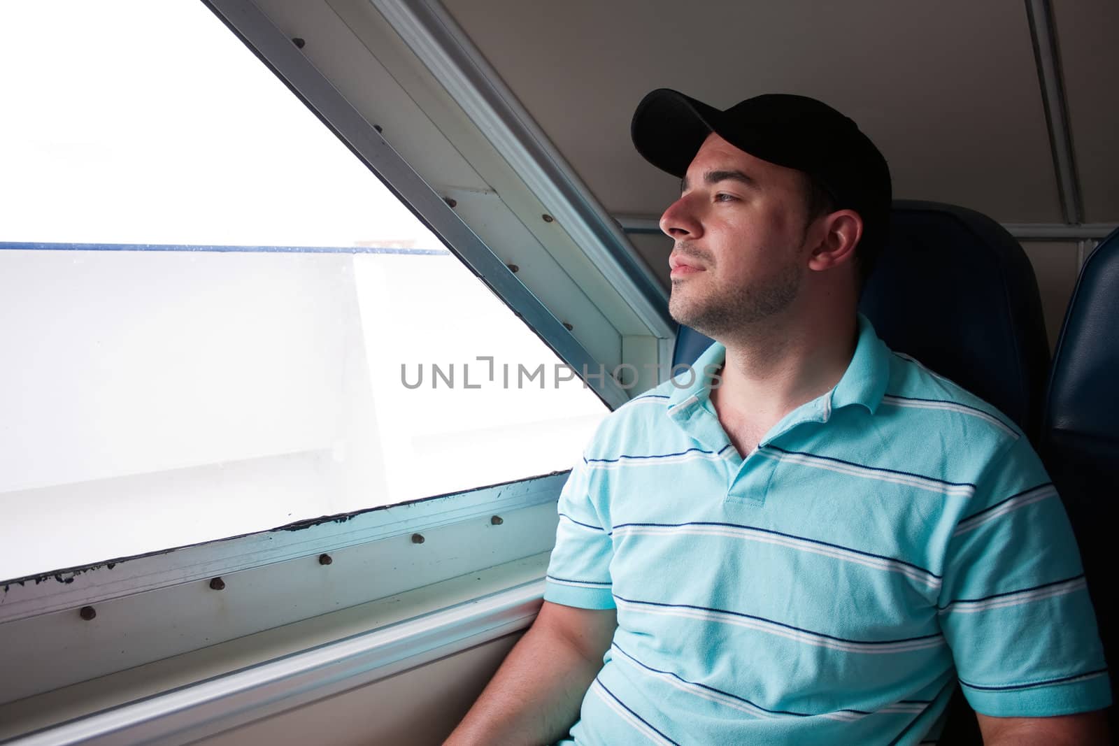 A tired looking passenger seated on a ferry boat looks out the window as he awaits arrival to his destination.