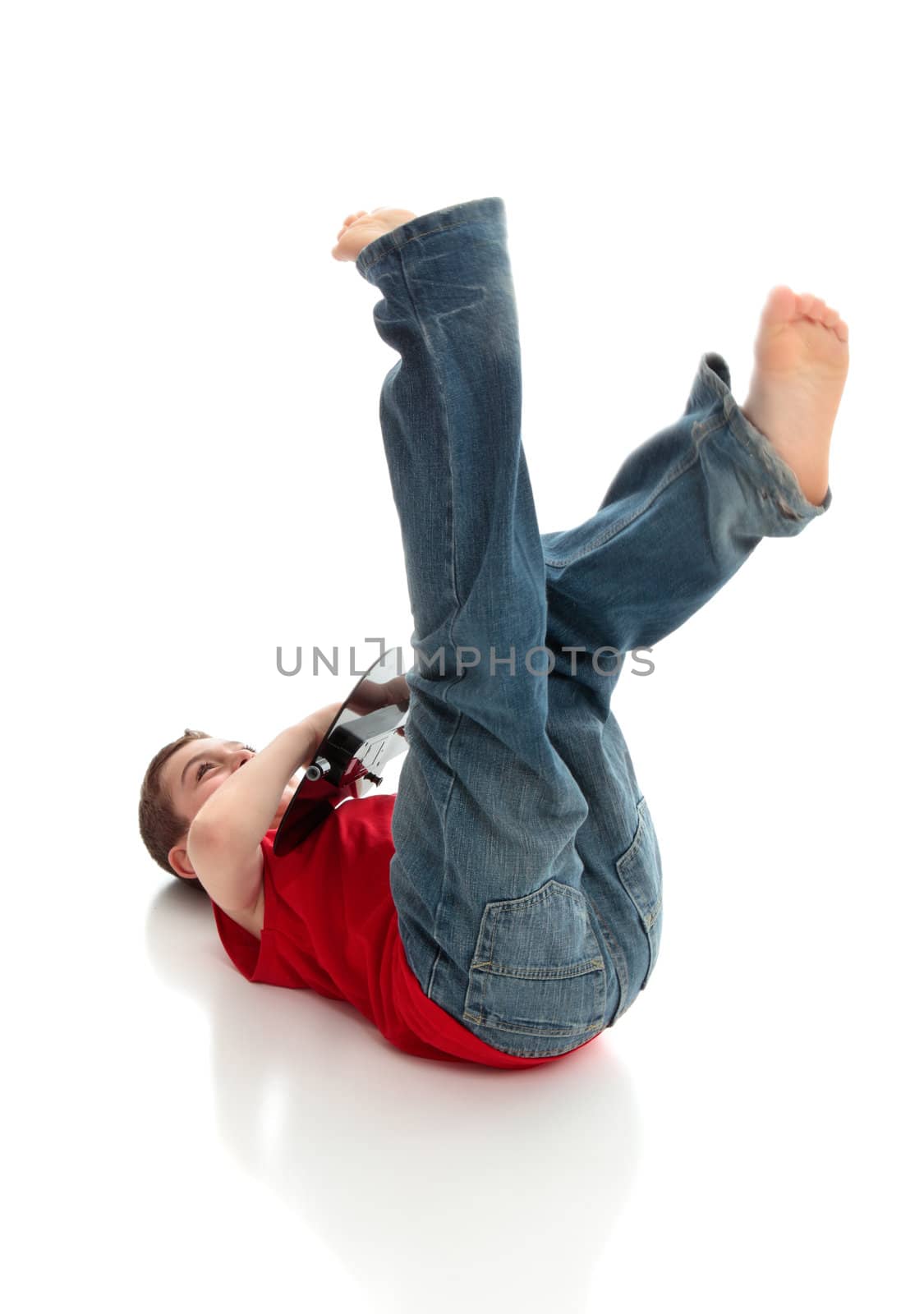 A little boy having fun playing a guitar.  He is lying on the ground with legs flailing in the air.  White background.