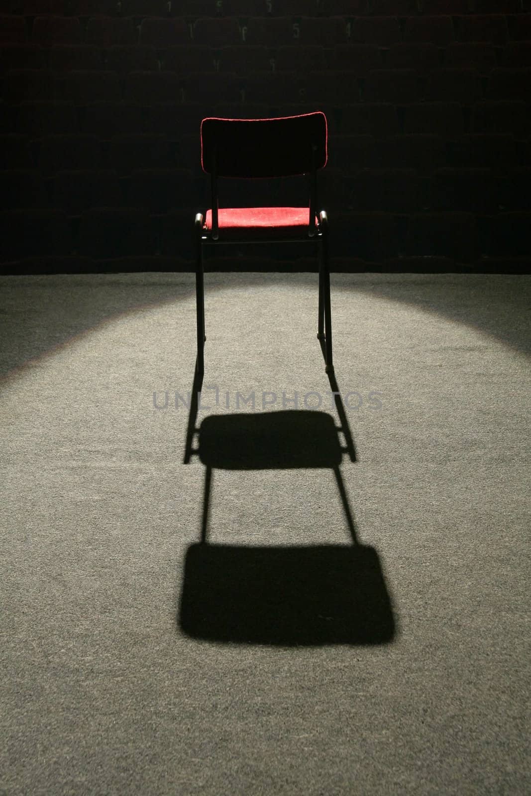 one red chair on stage, lighted with one spot light, empty seats in background