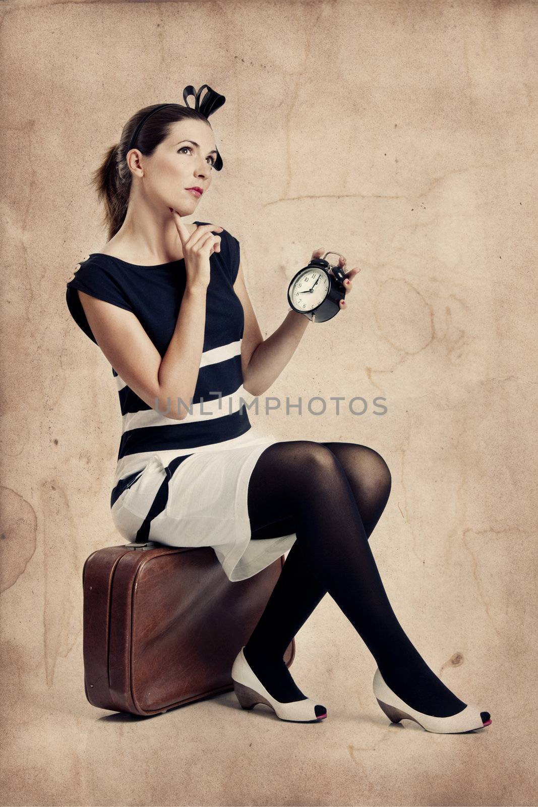 Beautiful young fashion woman sitting over a suitcase and holding a clock