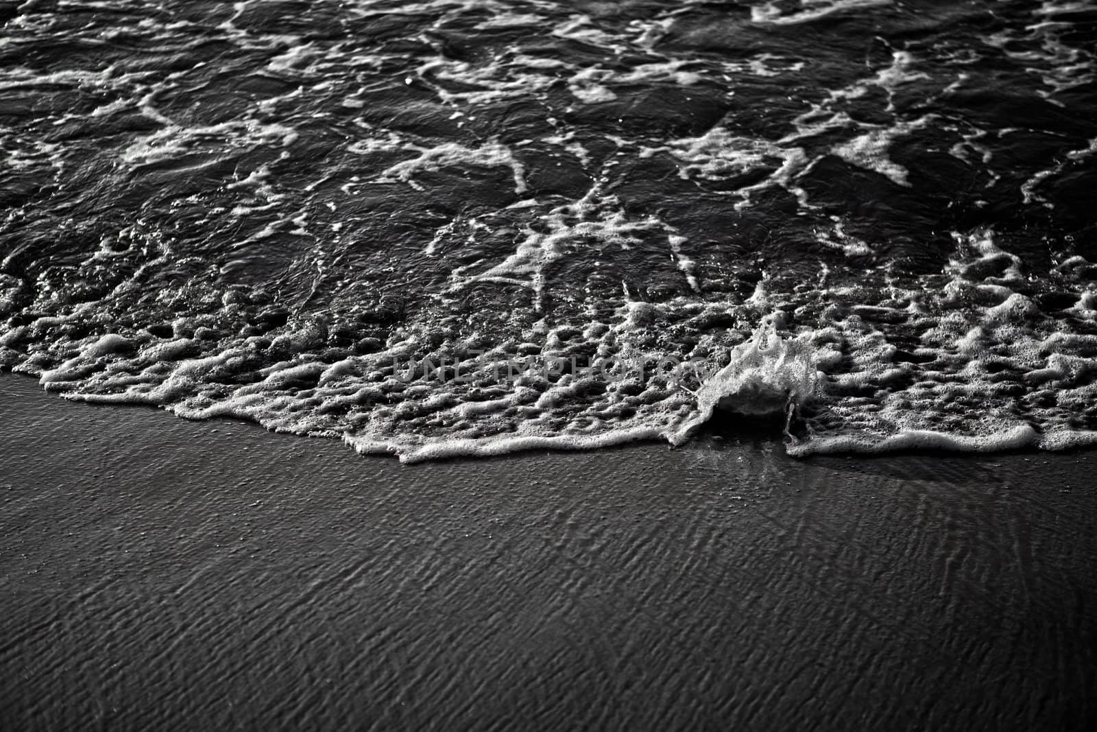 Monochrome image showing ocean water splashing onto a stone on a sandy beach