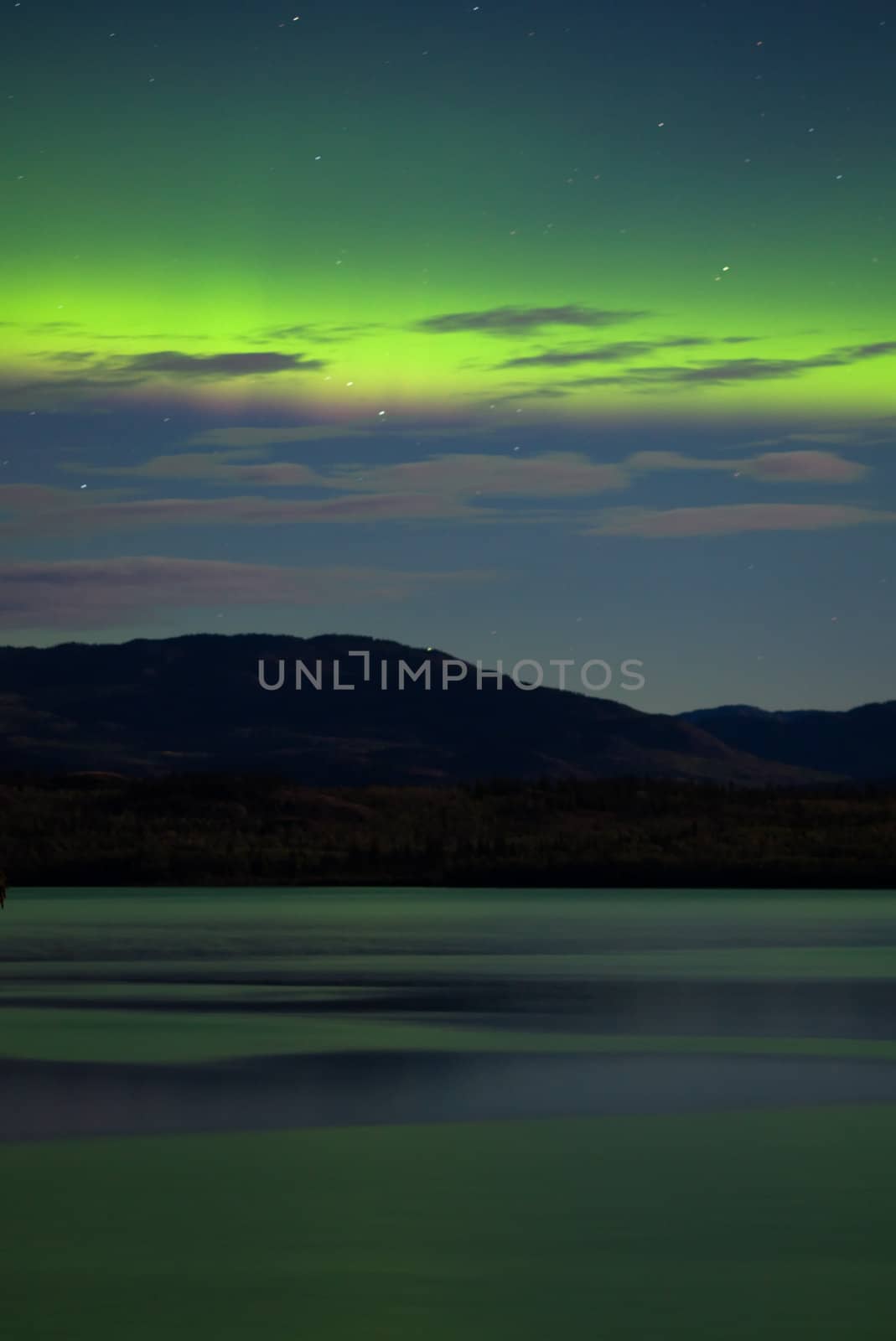 Intense Aurora borealis showing between clouds during moon lit night being mirrored on Lake Laberge, Yukon T., Canada.