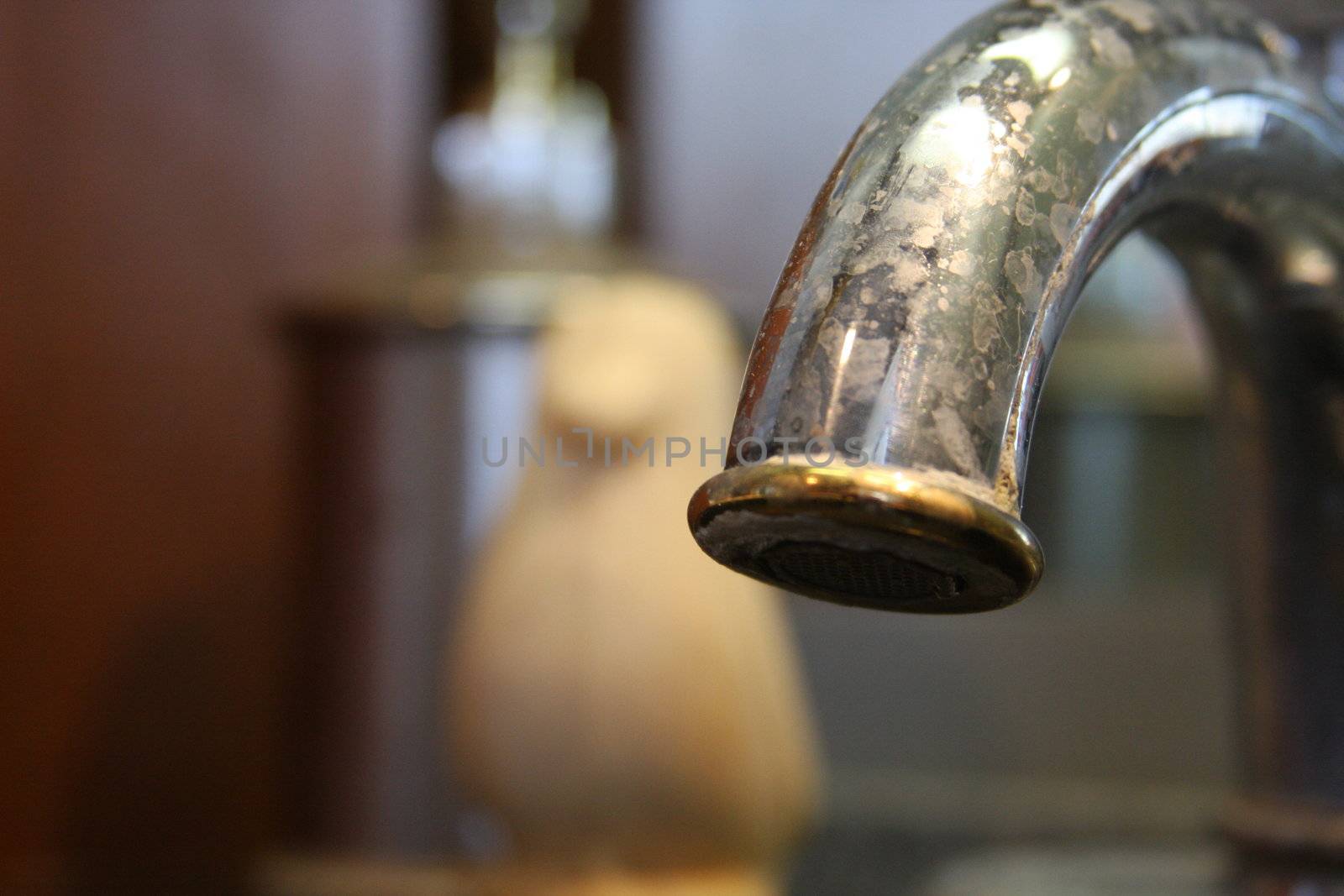 Focus on the end of a water-stained steel sink faucet with gold trim and a bit of mildew. An otherwise in good condition piece of hardware with some regular household grunge. Background shapes include metal soap dispenser and penguin-shaped bar soap.