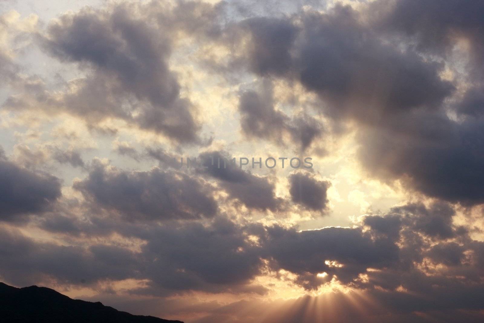 Sunset over a mountain, accentuated by storm clouds. Very clear and bright perspective with clouds layers that accentuate the horizon, while several sun rays penetrates the dark clouds. 