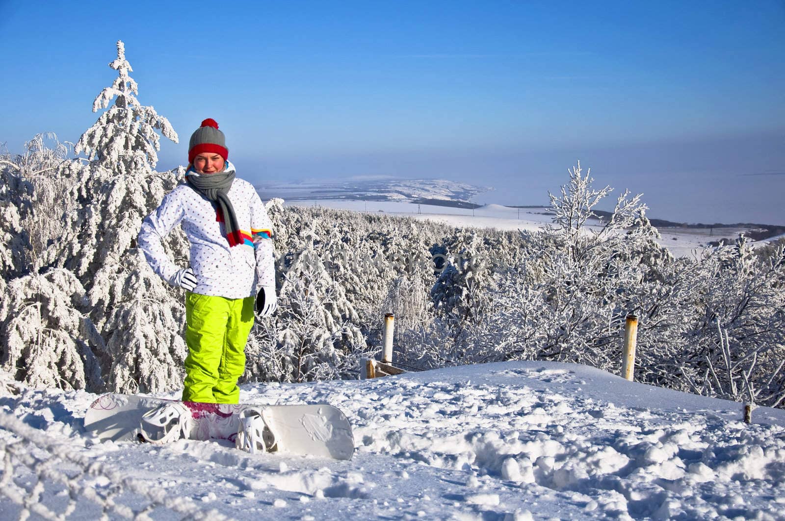 Beautiful woman with snowboard stands on top of the mountain. Against the backdrop of snow-covered pine trees and blue sky.