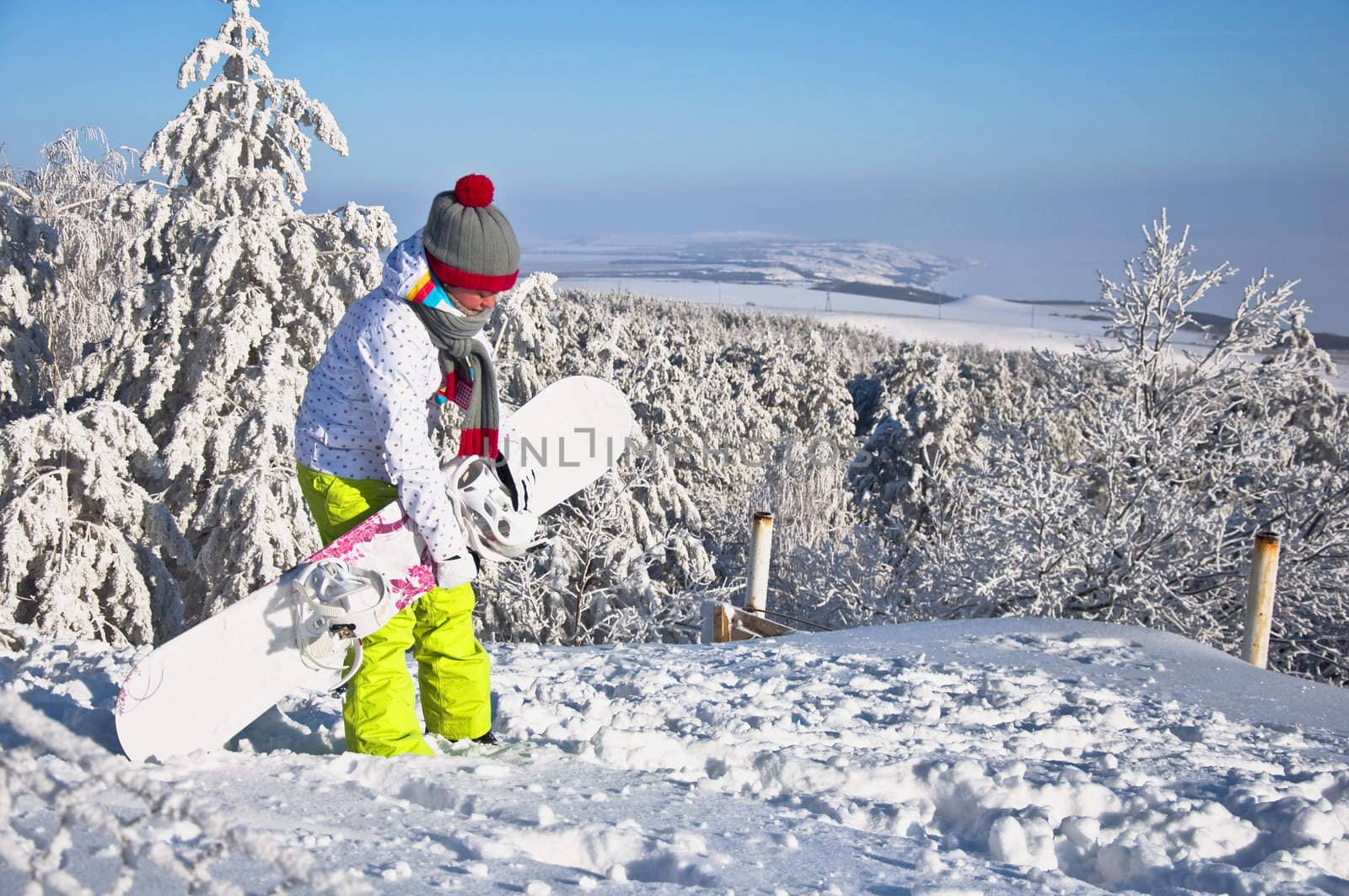 Beautiful woman with snowboard stands on top of the mountain. Against the backdrop of snow-covered pine trees and blue sky.