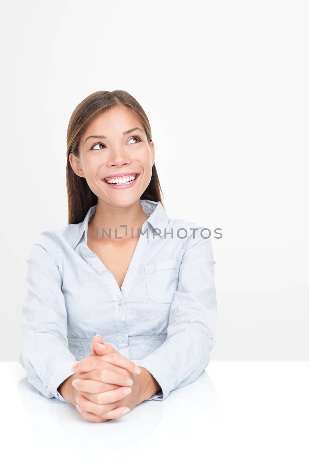 Woman smiling looking up thinking sitting at table. Positive, optimistic and friendly looking Asian / Caucasian businesswoman isolated on white background.