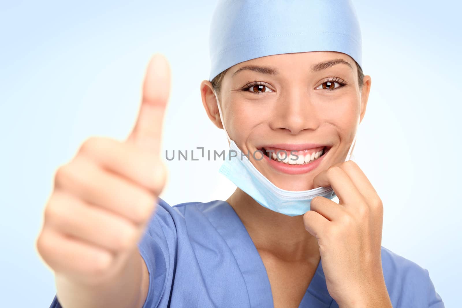 Young doctor or nurse giving thumbs up taking off the surgeon mask smiling. Asian / Caucasian female model in her mid 20s.