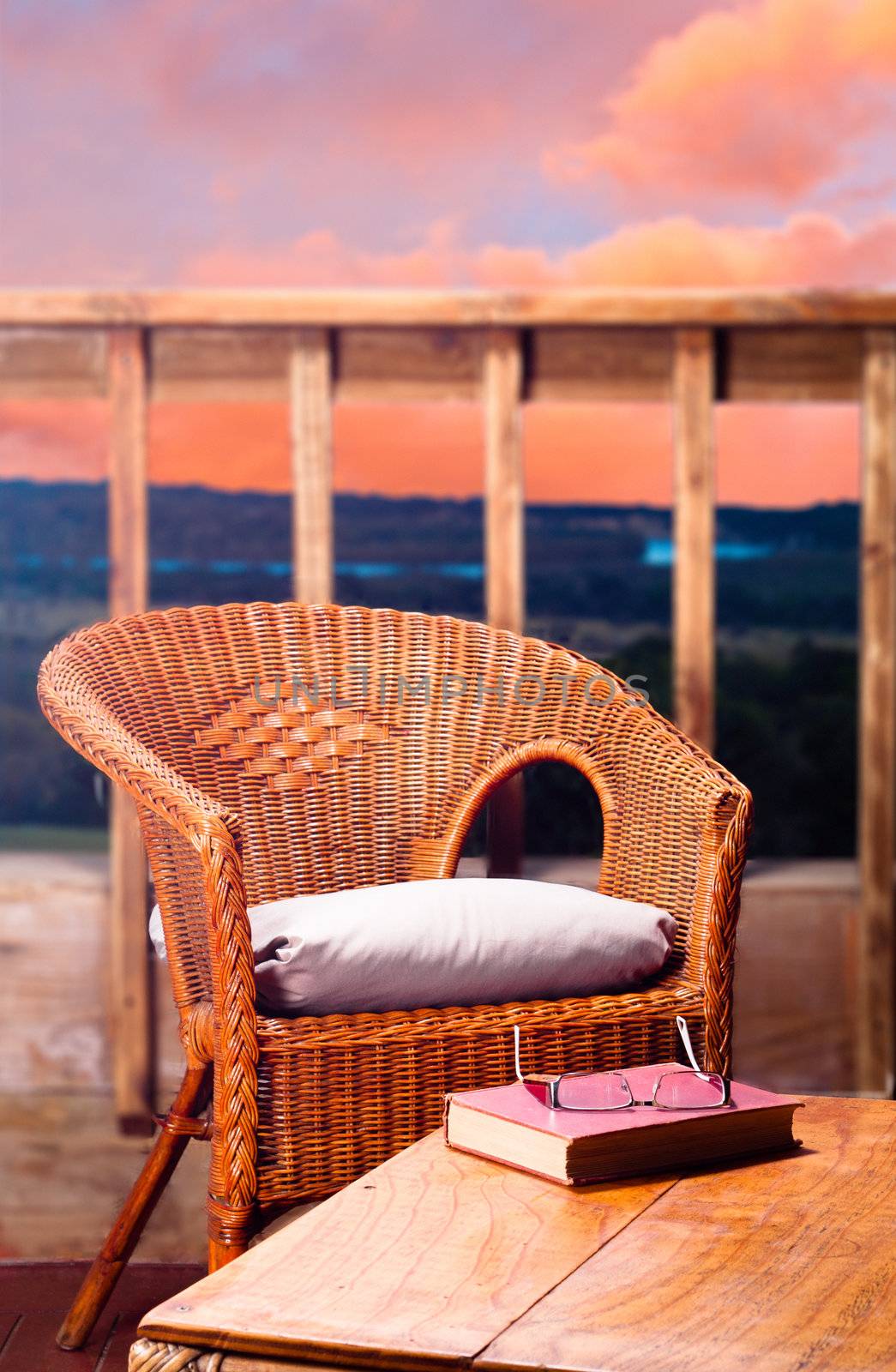 Wicker chair with pillow next to a wooden table with a book and reading glasses on it, with a view of a river at sunset in the background