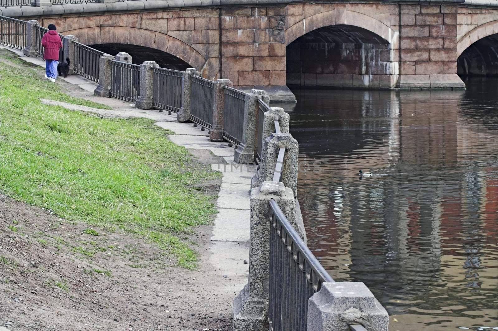 River Embankment, metal railings, and senior woman with dog in remote distance.