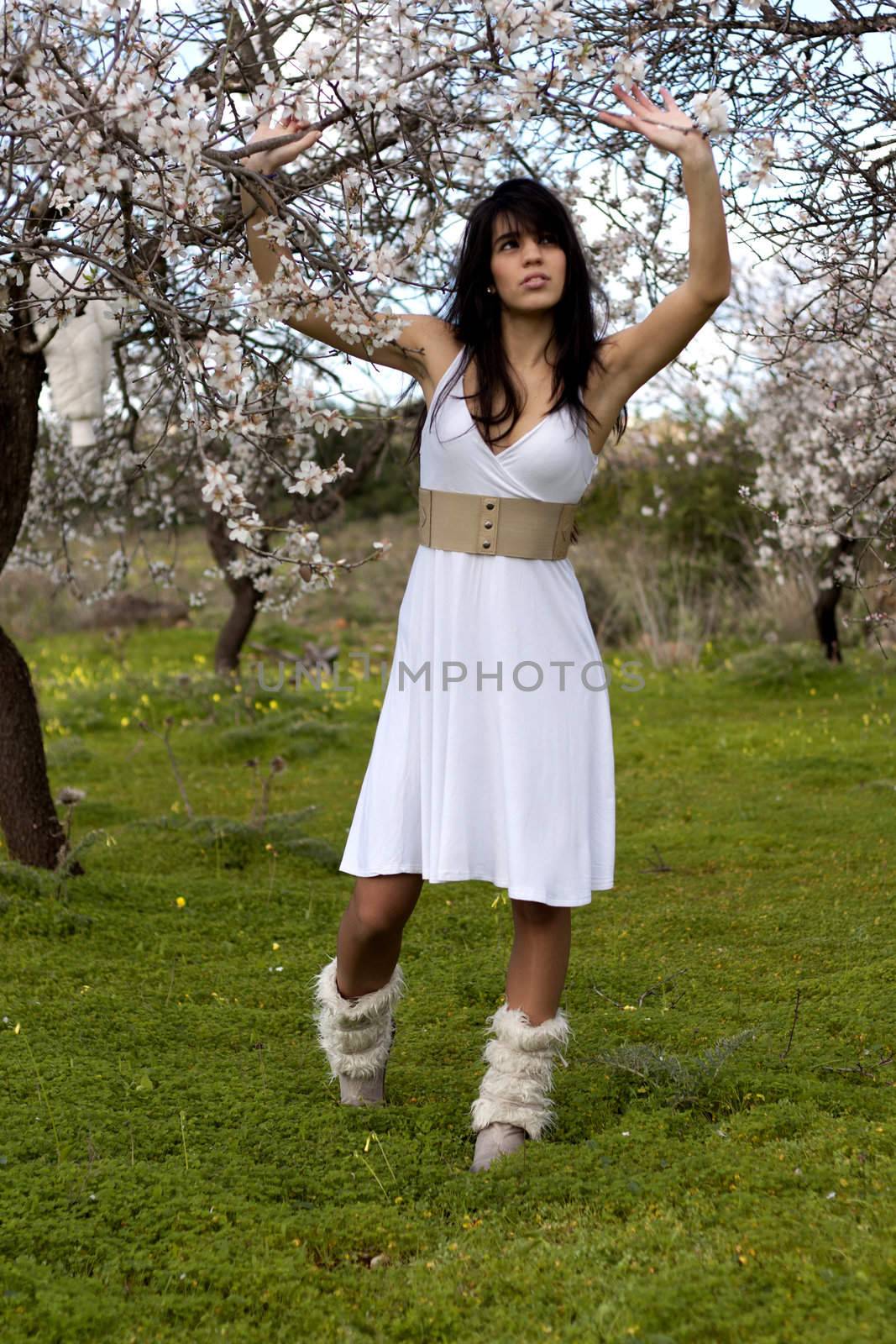 View of a beautiful girl on a white dress on a green grass field next to a almond tree