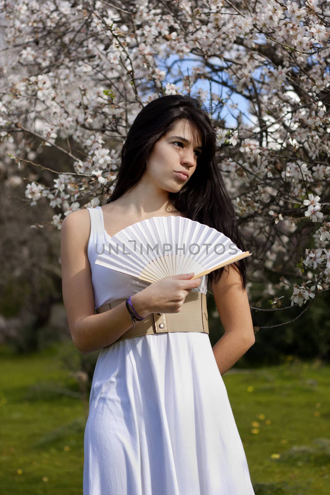 View of a beautiful girl on a white dress on a green grass field next to a almond tree