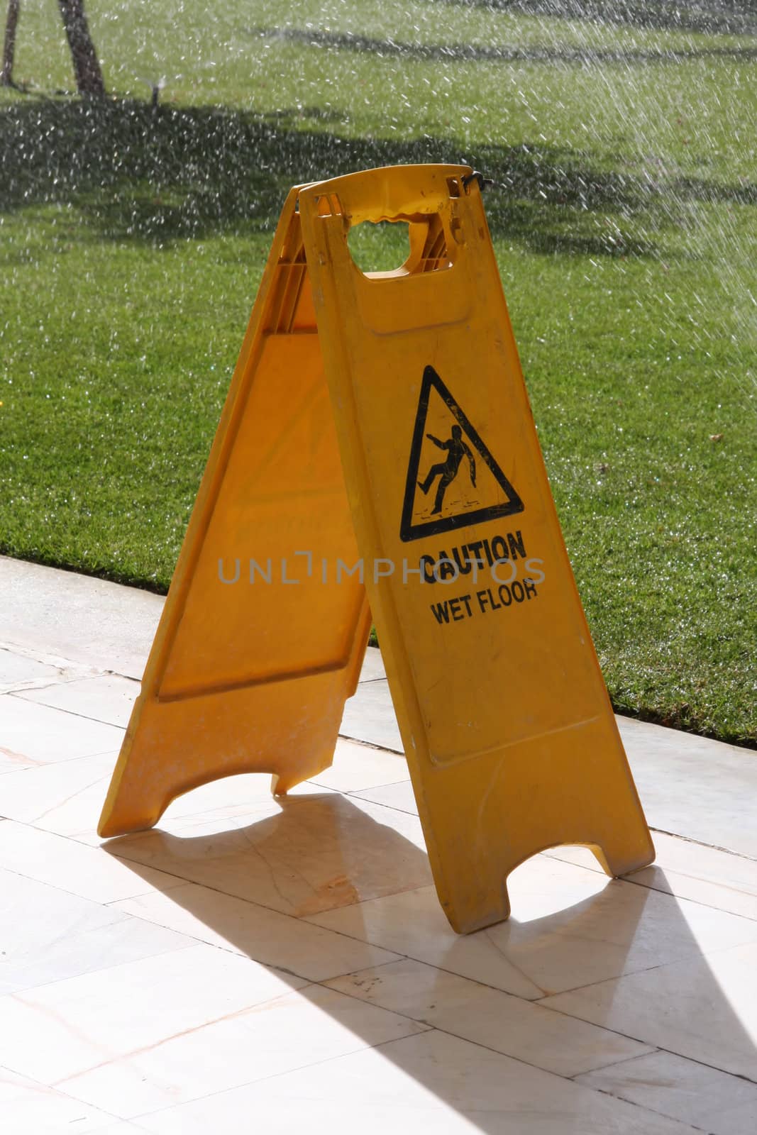 Yellow cleaning sign, caution wet floor, sprinklers on in the background