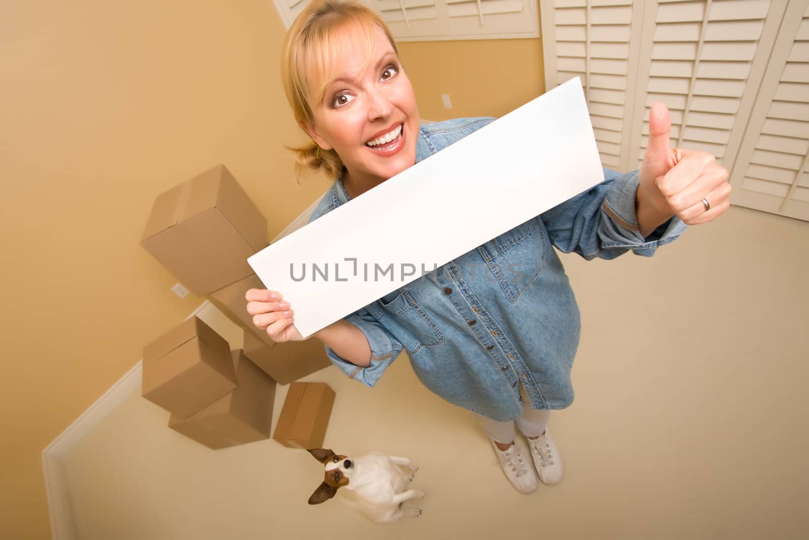 Excited Woman with Thumbs Up and Doggy Holding Blank Sign Near Moving Boxes in Empty Room Taken with Extreme Wide Angle Lens.

