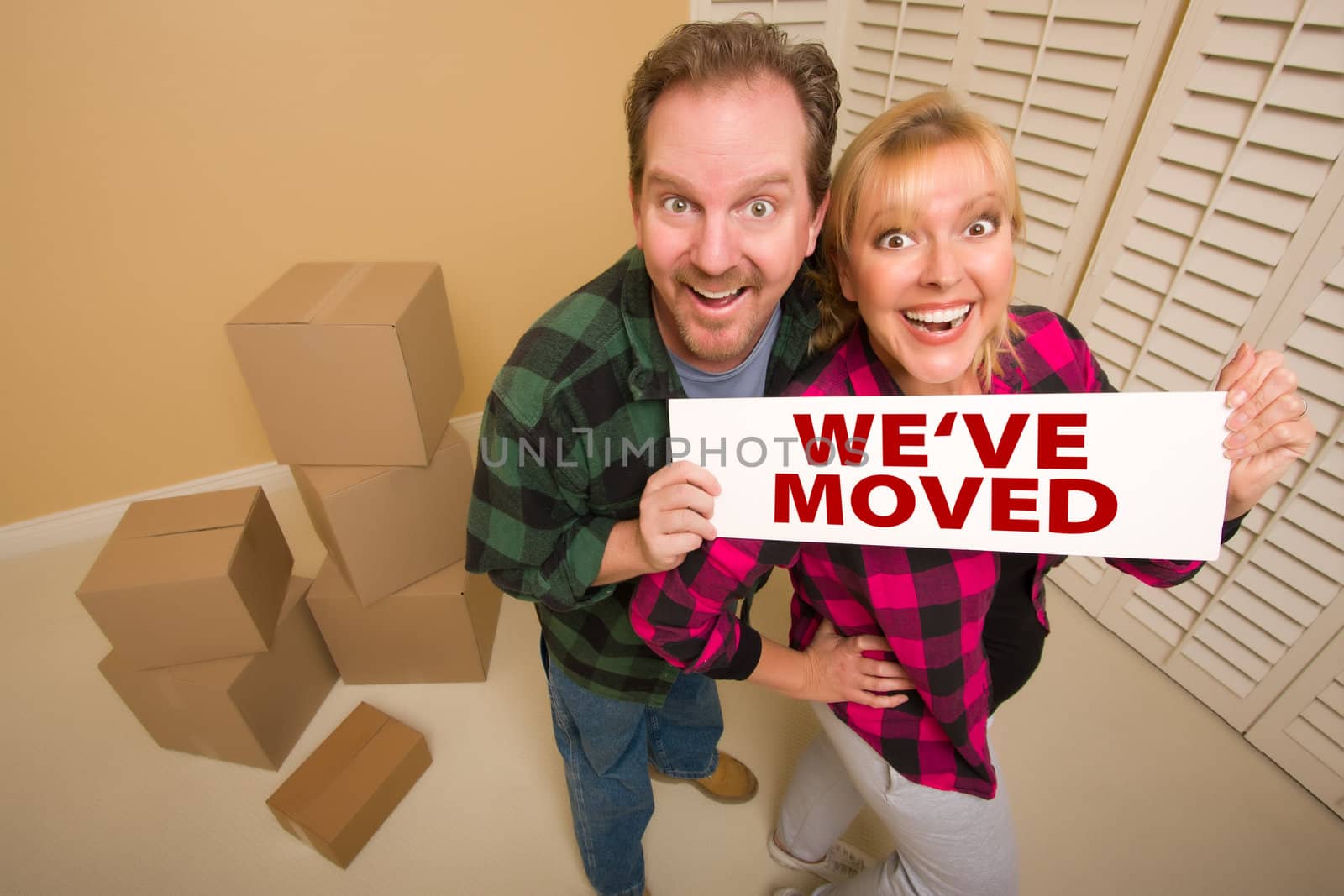 Goofy Couple Holding We've Moved Sign in Room with Packed Cardboard Boxes.