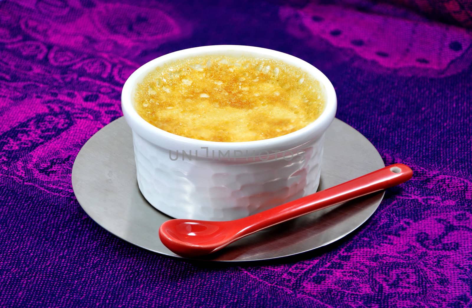 Home-made custard with a red ceramic spoon and an iron steel saucer on a purple tablecloth