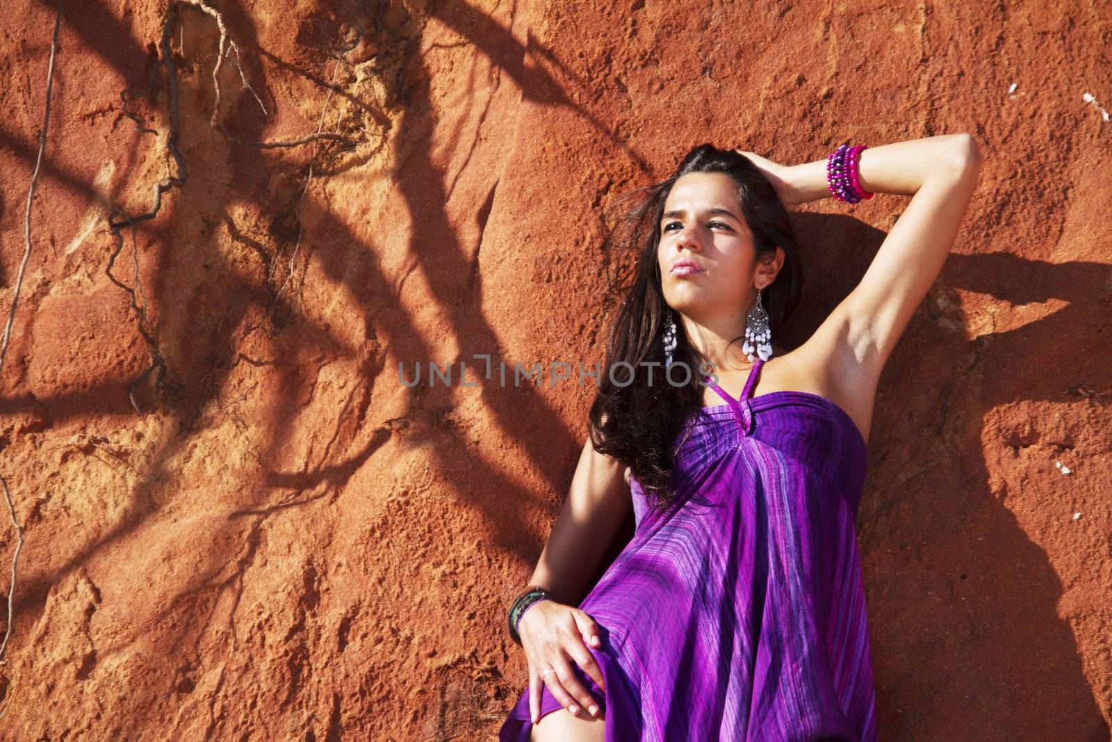 View of a beautiful young girl posing with a purple dress next to a cliff on the beach. 