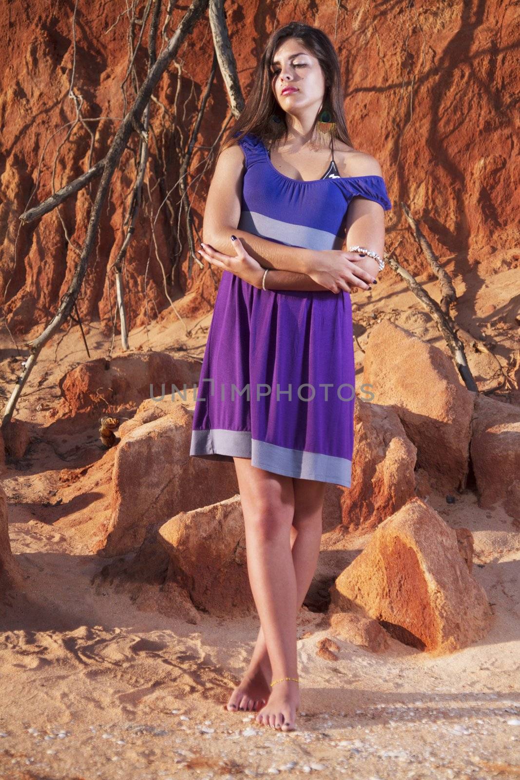 View of a beautiful young girl with a purple dress standing on a red cliff on the beach.