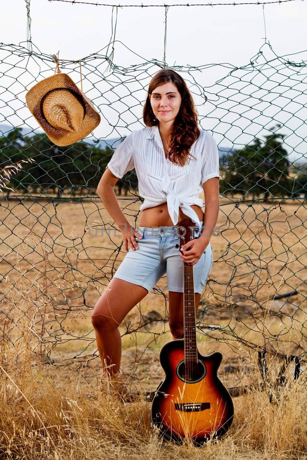 View of a beautiful young country girl with a guitar on the fence of the grassland. 