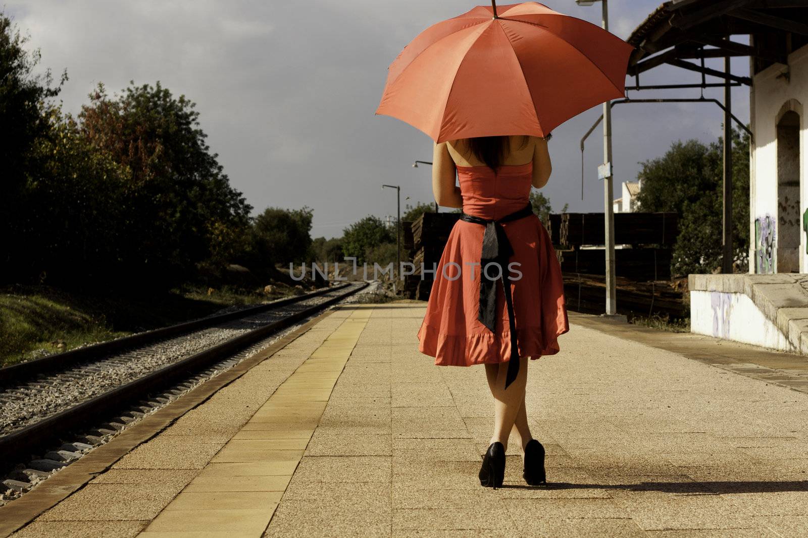 View of a beautiful woman with red dress and umbrella on a train station.