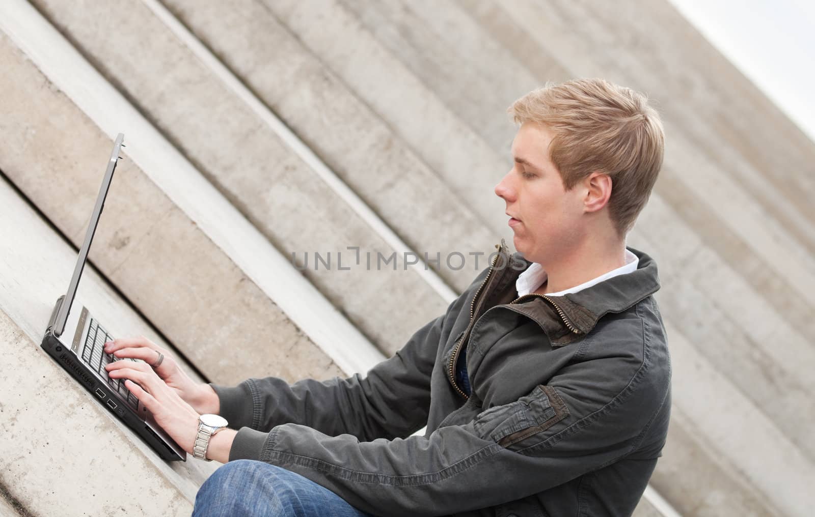 Young blond handsome smiling guy sitting outdoors with laptop computer