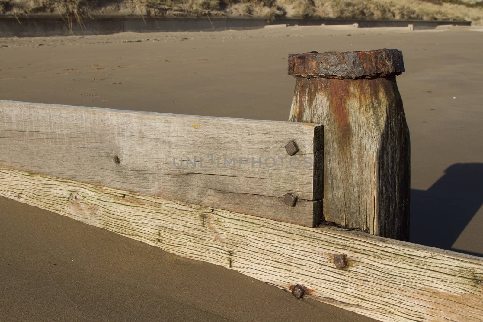 Wooden planks, water smoothed, attached to a post with metal bolts form a sand barrier on a beach.