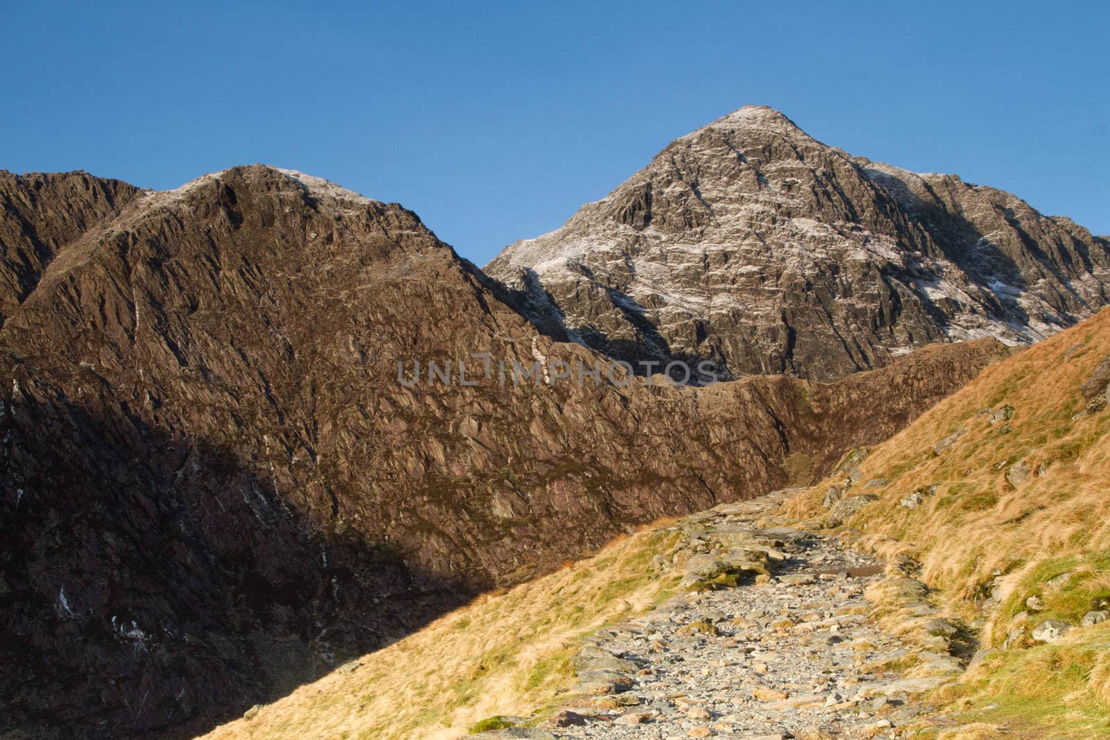 A footpath leads up past a grassy bank towards large mountainous cliffs with ridgelines and peaks.