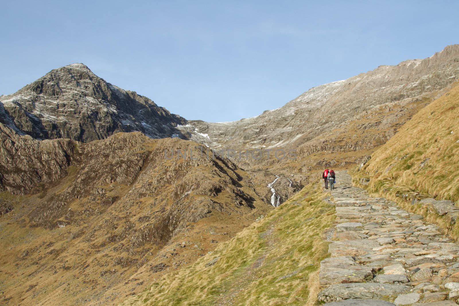 Two people walk up a stone footpath towards a high rocky peak.